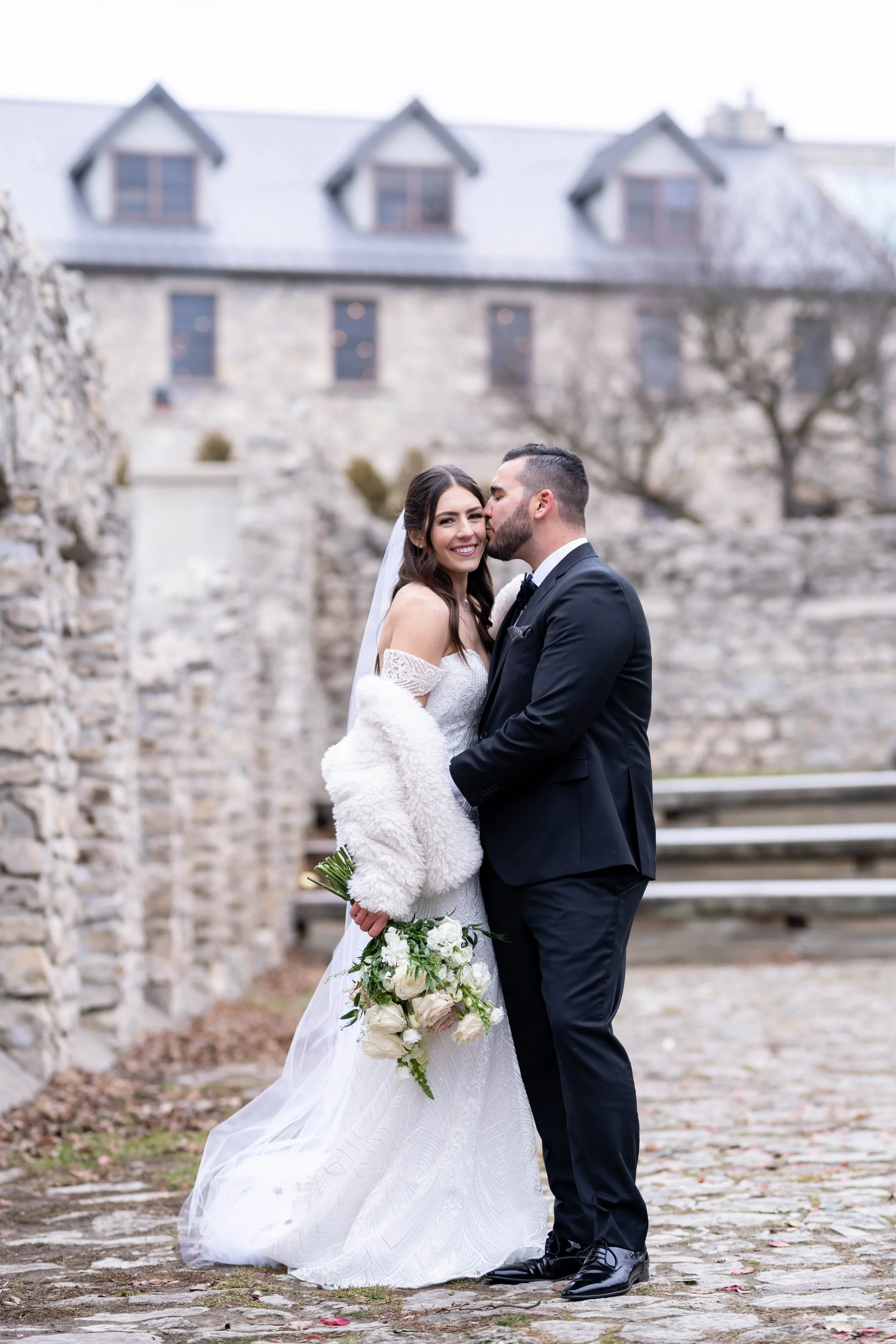 A bride and groom standing outdoors, smiling and sharing a kiss in front of an old stone building with a rocky wall and leafless trees in the background. The bride holds a bouquet of white flowers and wears a white lace wedding dress with off-the-sho