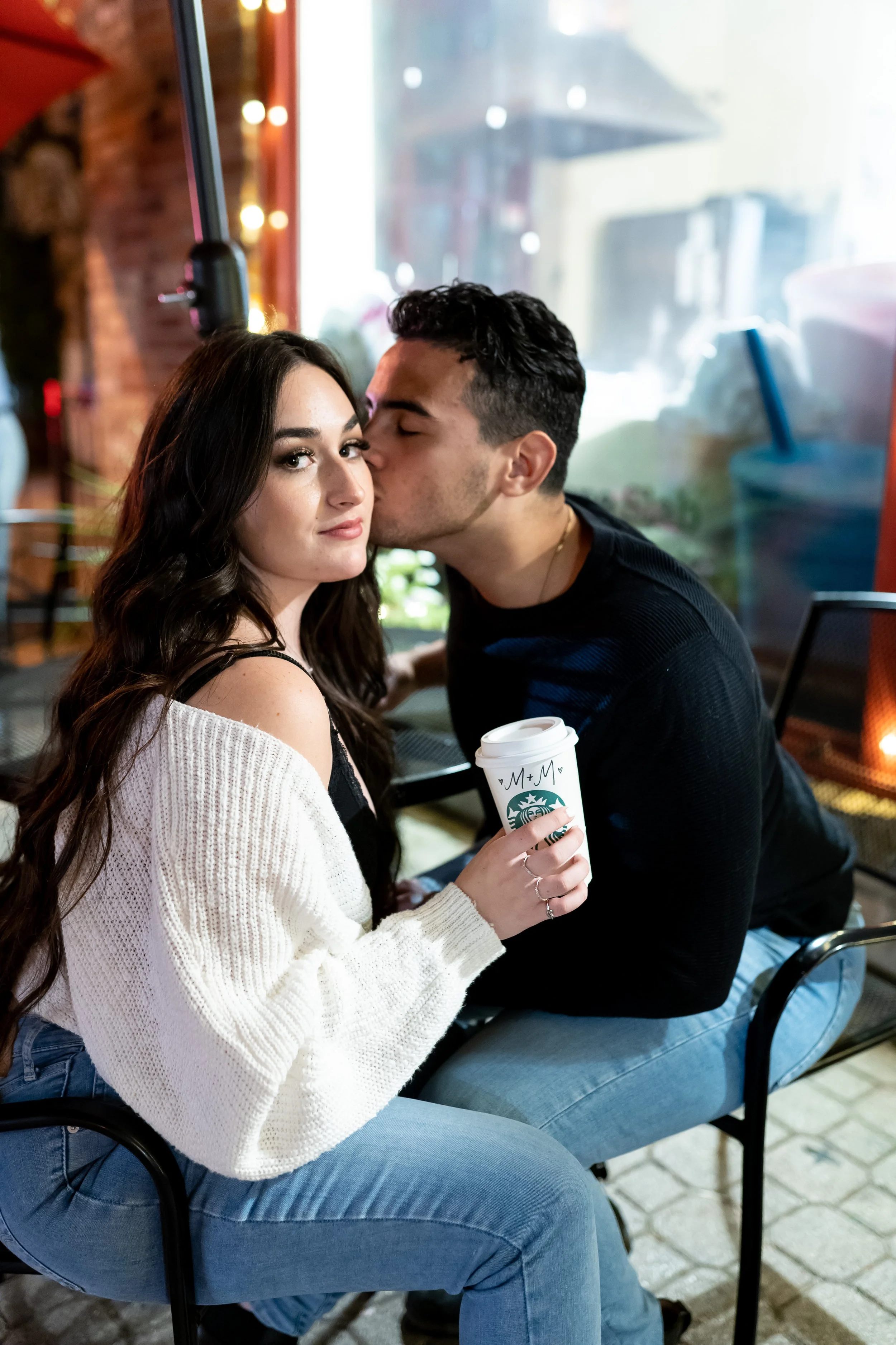 A young woman with long brown hair, wearing a white sweater, holding a Starbucks coffee cup, sitting on a chair inside a coffee shop. A young man with dark curly hair, wearing a black shirt, is kissing her on the cheek. The background shows a large w