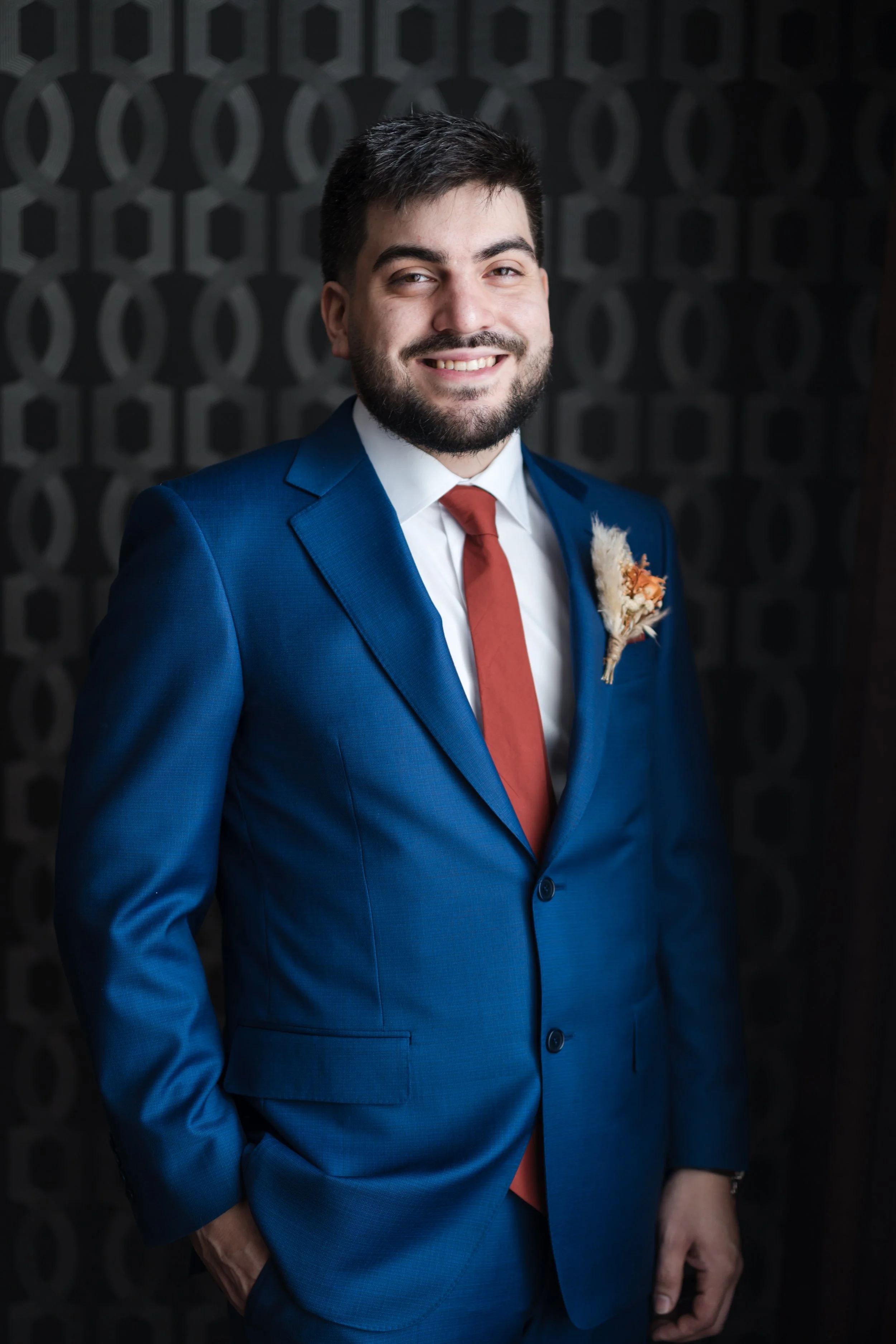 A man in a blue suit with a white shirt and red tie, smiling and standing against a dark patterned background.