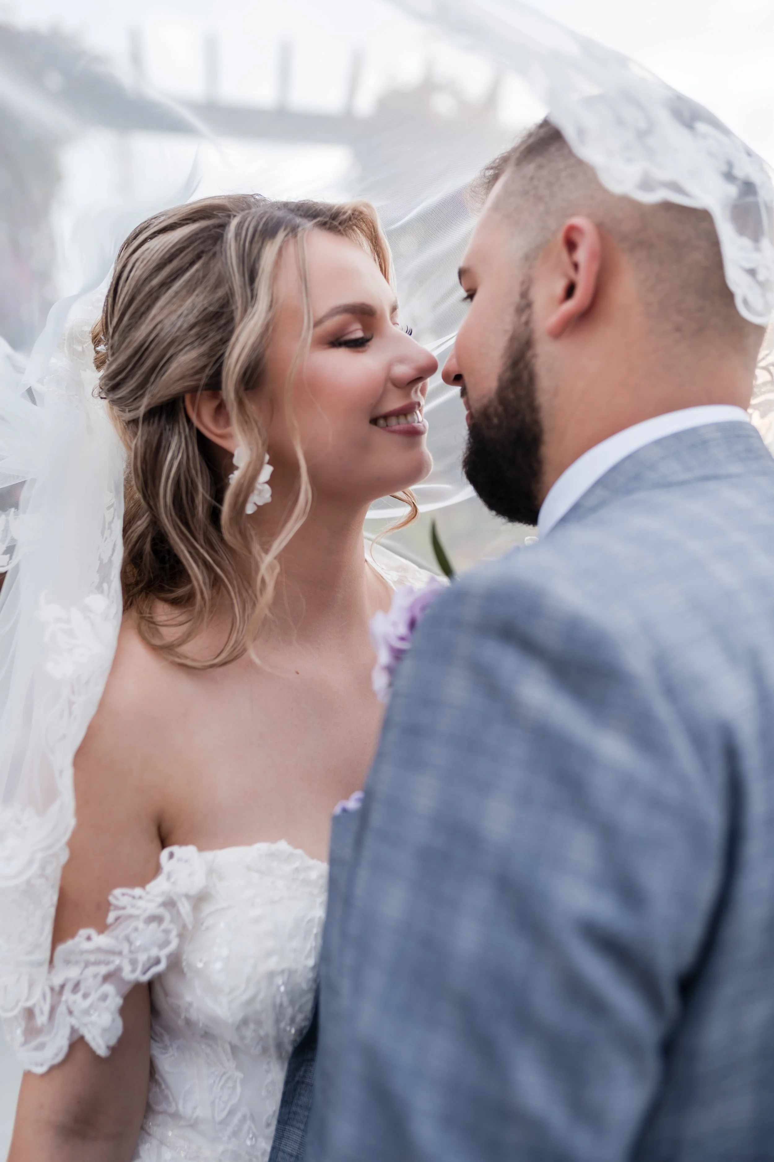 A bride and groom face each other, smiling with their noses nearly touching, under a lace veil in a close-up shot.