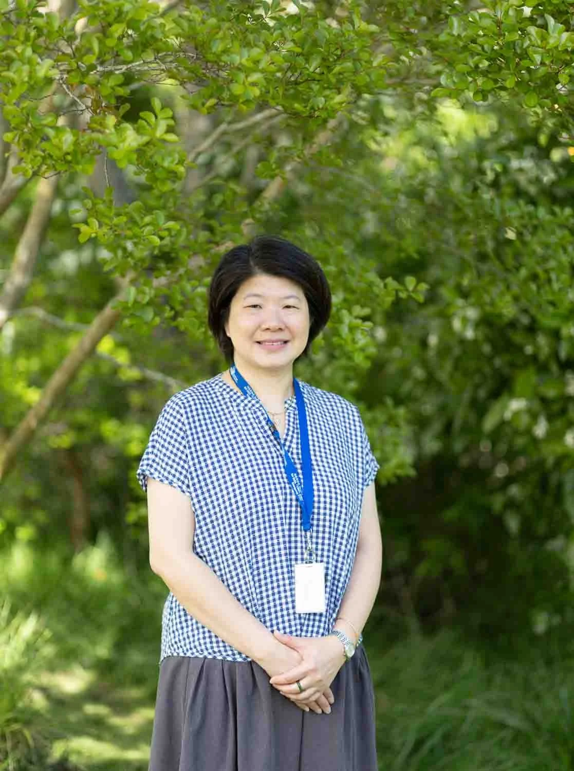 A woman with short black hair smiling outdoors, standing in front of green trees, wearing a blue and white checkered blouse, a gray skirt, and a blue lanyard with an ID badge.