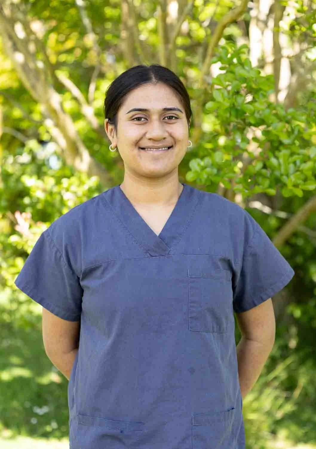A woman wearing blue medical scrubs standing outdoors in front of green trees and bushes, smiling at the camera.