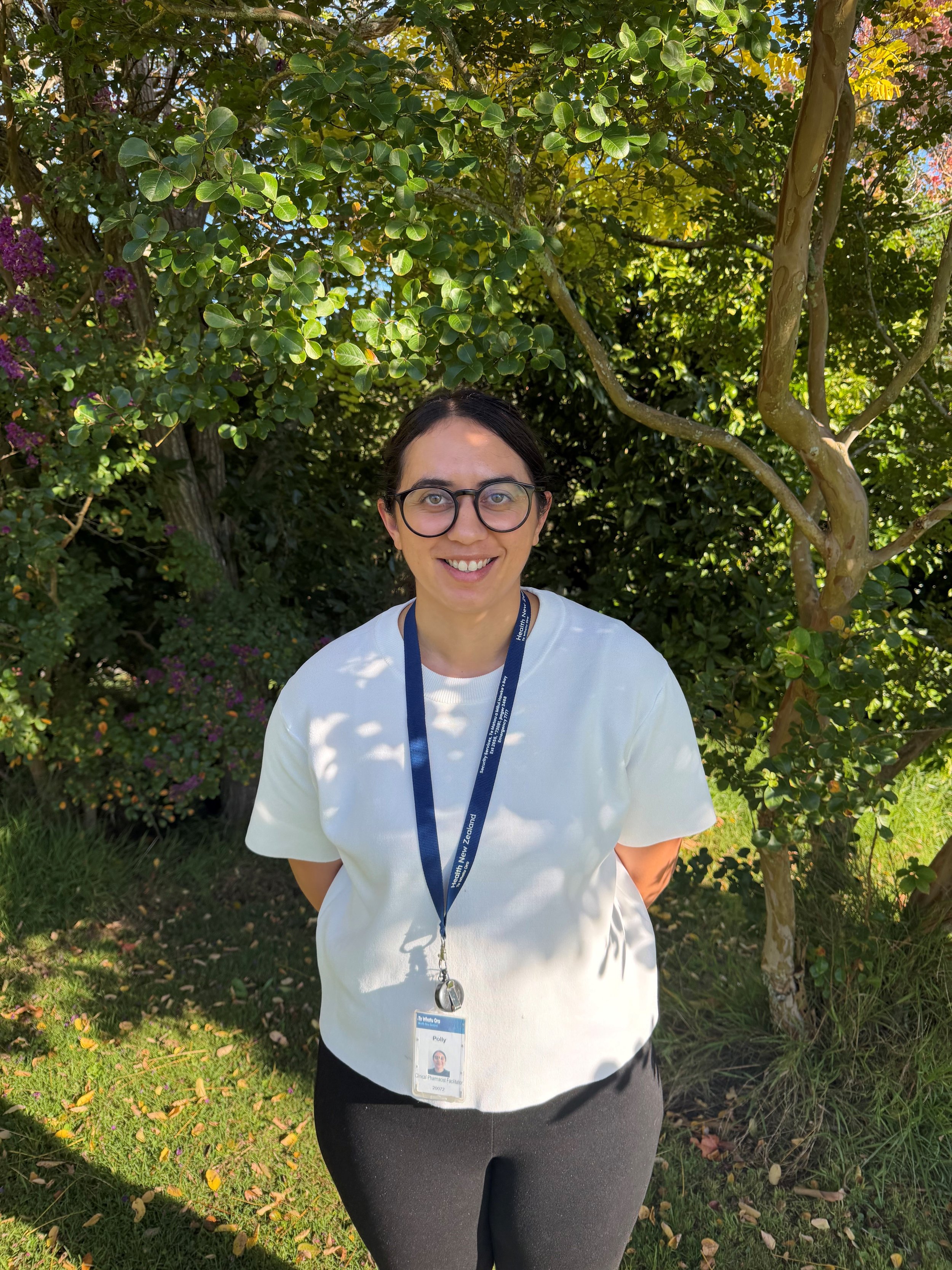 A woman with short black hair smiling outdoors, standing in front of green trees, wearing a blue and white checkered blouse, a gray skirt, and a blue lanyard with an ID badge.