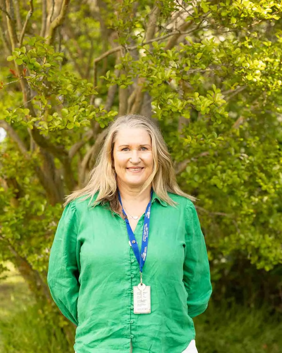 A woman standing outdoors in front of leafy green trees, smiling, wearing a green blouse and a lanyard with an ID badge.