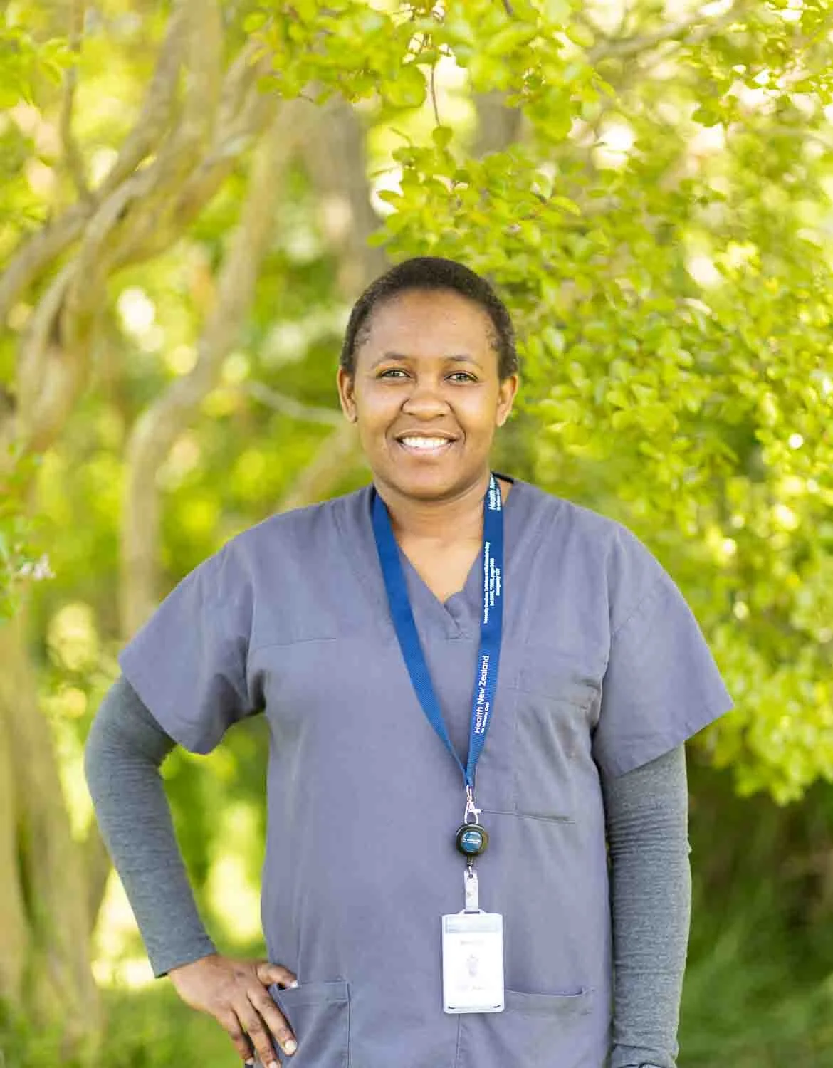 A smiling woman in gray scrubs outdoors in a green, leafy setting, wearing a lanyard with ID badge.