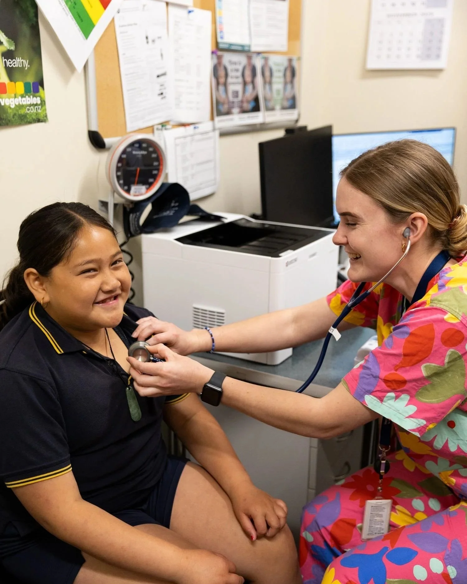 A young girl sitting in a medical examination room smiling while a nurse uses a stethoscope on her chest.