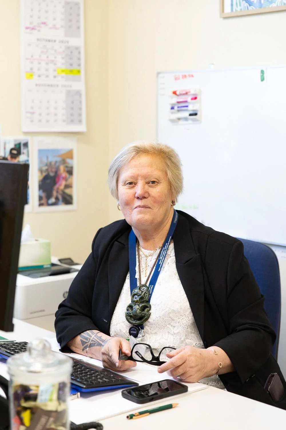 An older woman with short blonde hair sitting at a desk in an office, holding glasses in her hand, with a computer, a phone, and office supplies on the desk, and a whiteboard and calendar on the wall behind her.