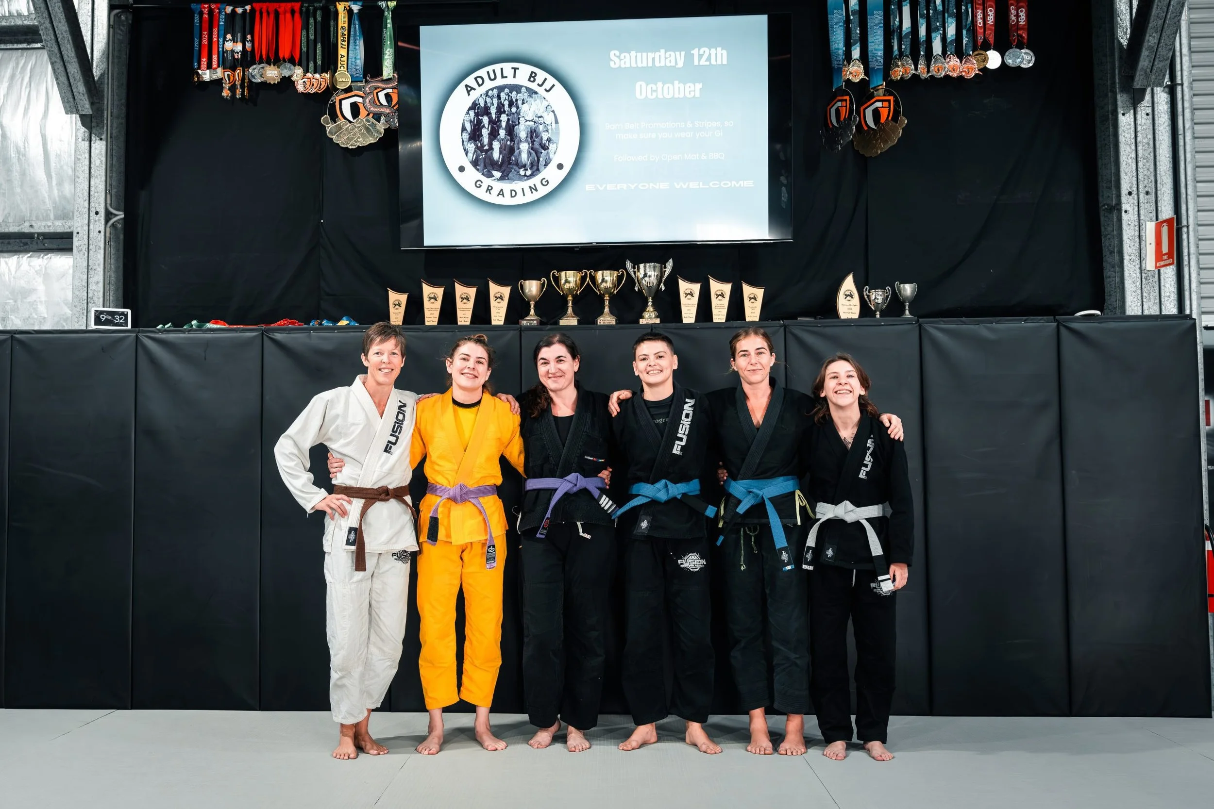 Group of six women in Brazilian Jiu-Jitsu gis standing together in a dojo, smiling and posing for a photo. Behind them is a black wall with a digital screen displaying information about an event, medals, trophies, and awards on a table.