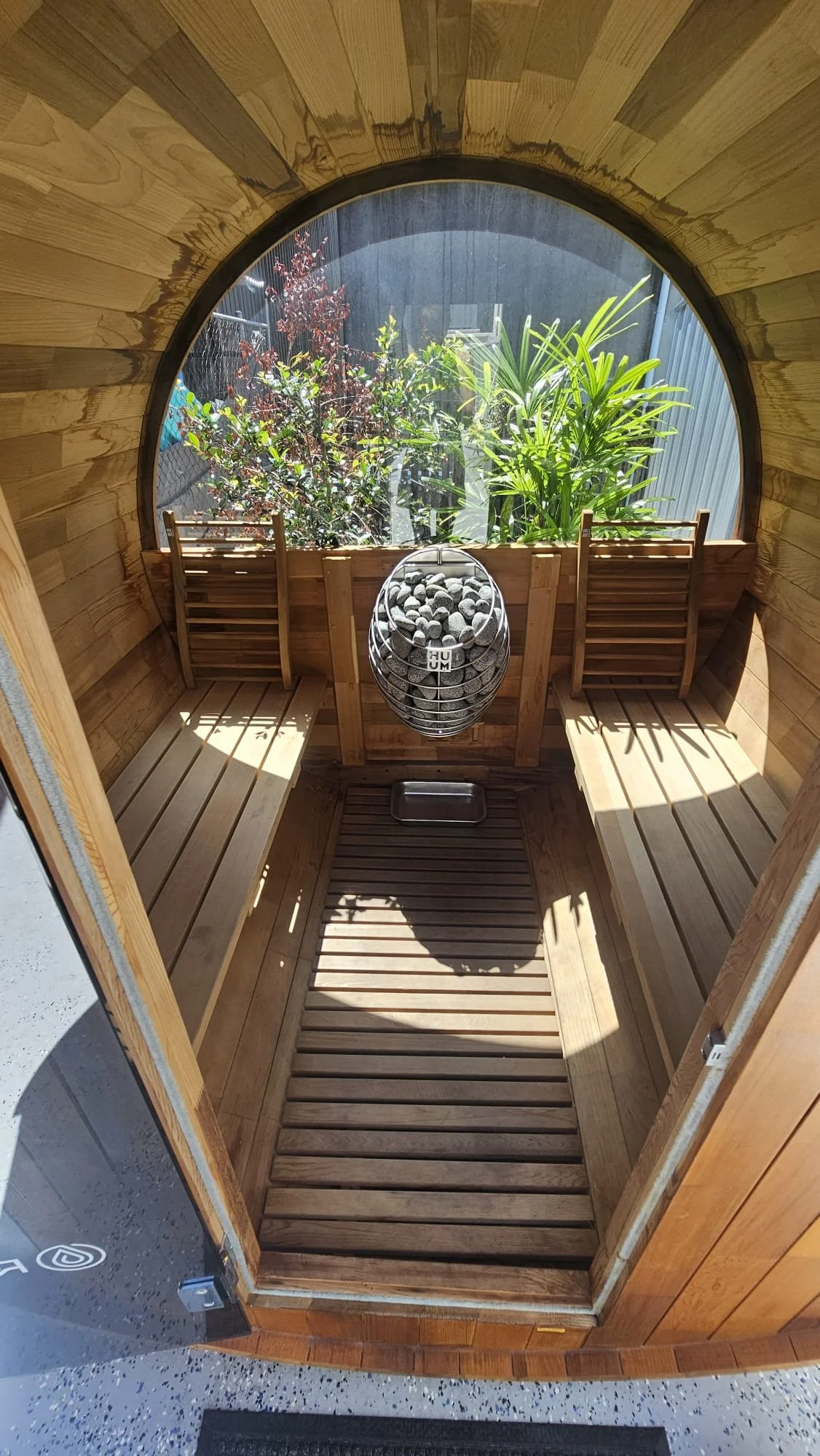 Inside a wooden sauna with benches on each side and a window revealing green plants outside. A hanging heater with stones is in the center.