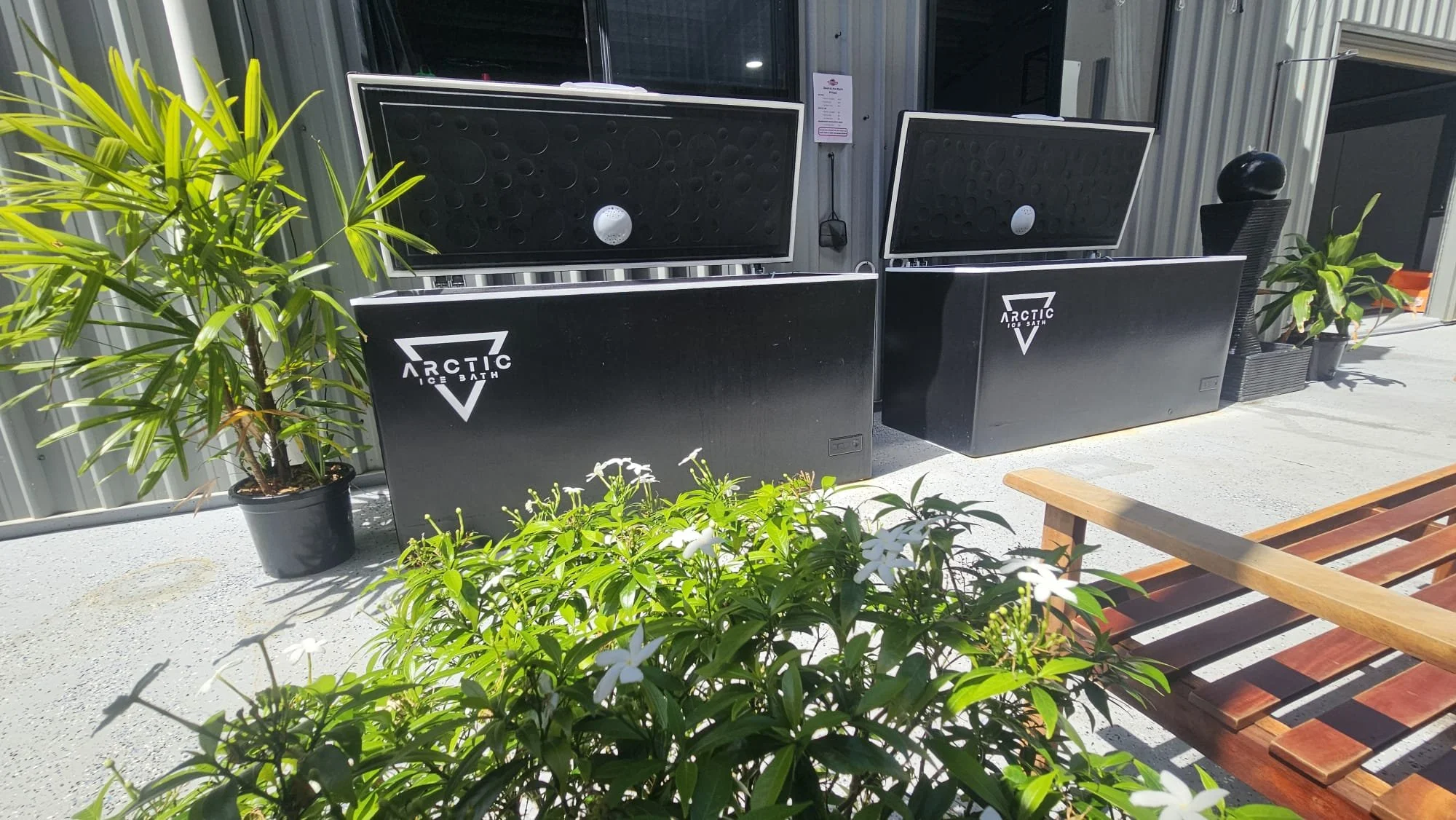 Outdoor scene with two black ice bath tubs labeled 'Arctic Ice Bath,' surrounded by plants and a wooden bench, outside a building with metal siding.