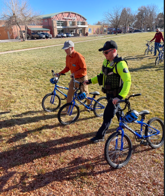 Two men with bicycles standing on grass, with a building and other people riding bikes in the background on a sunny day.