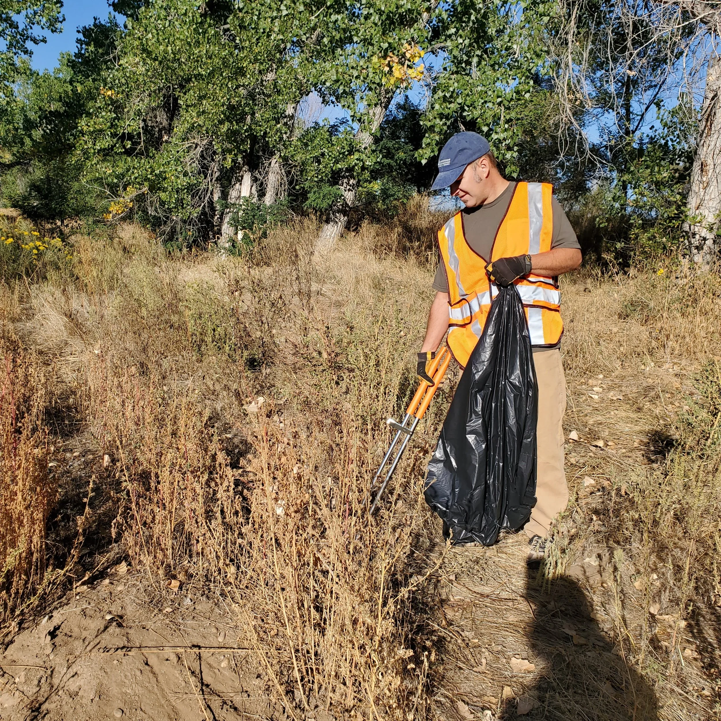 A man wearing an orange safety vest, beige pants, and a blue cap, using trash pick-up tools and a black garbage bag to clean up a dry, grassy outdoor area with trees in the background on a sunny day.