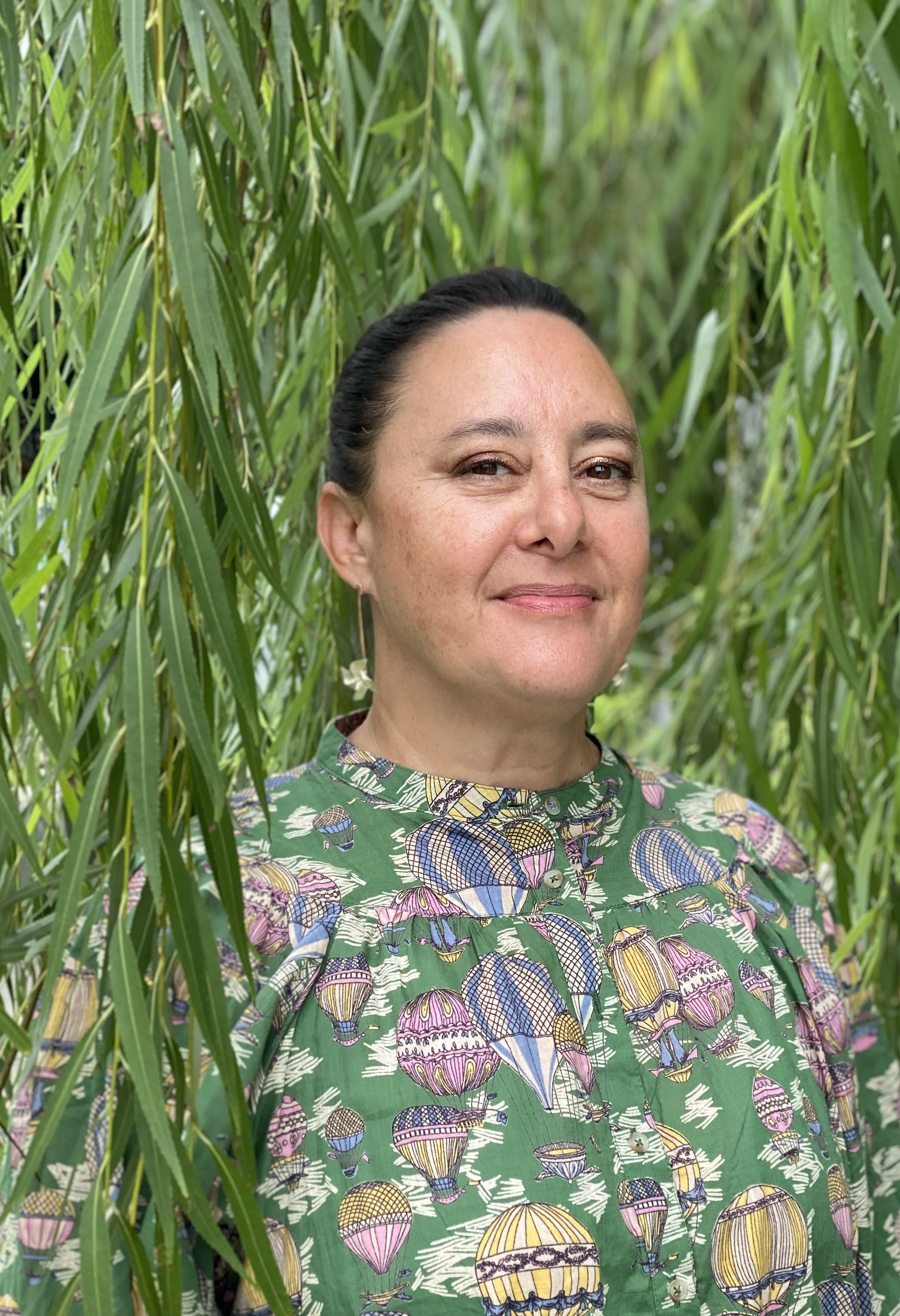 A woman with dark hair tied back, wearing a colorful shirt with hot air balloon patterns, standing amidst tall green grass or reeds.