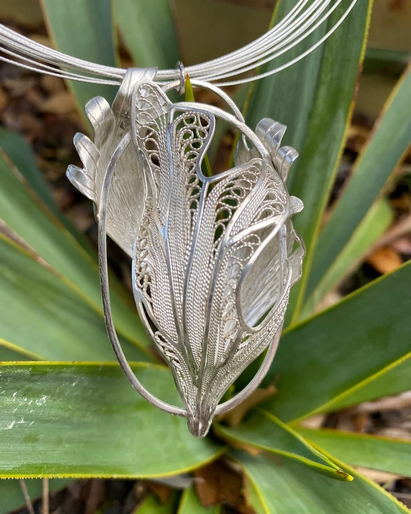 Close-up of a silver jewelry piece with intricate leaf and feather designs, hanging on a green plant background.