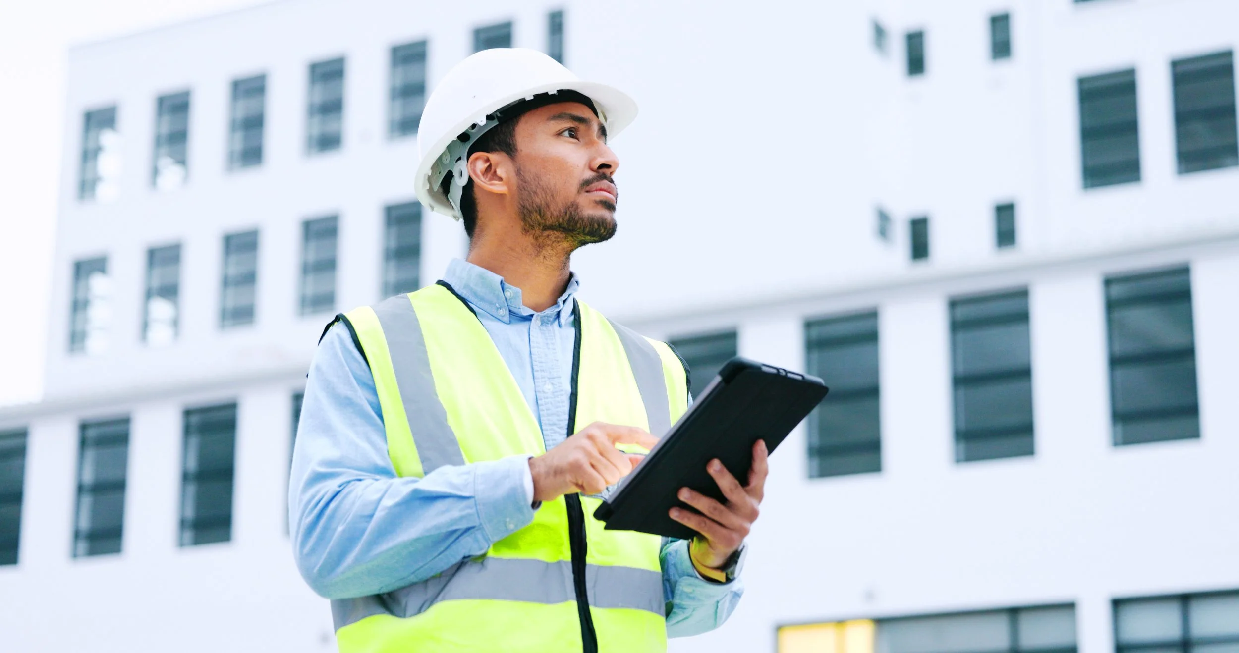 A man with a white hardhat, blue long sleeve button up, and yellow reflective vest holding a tablet