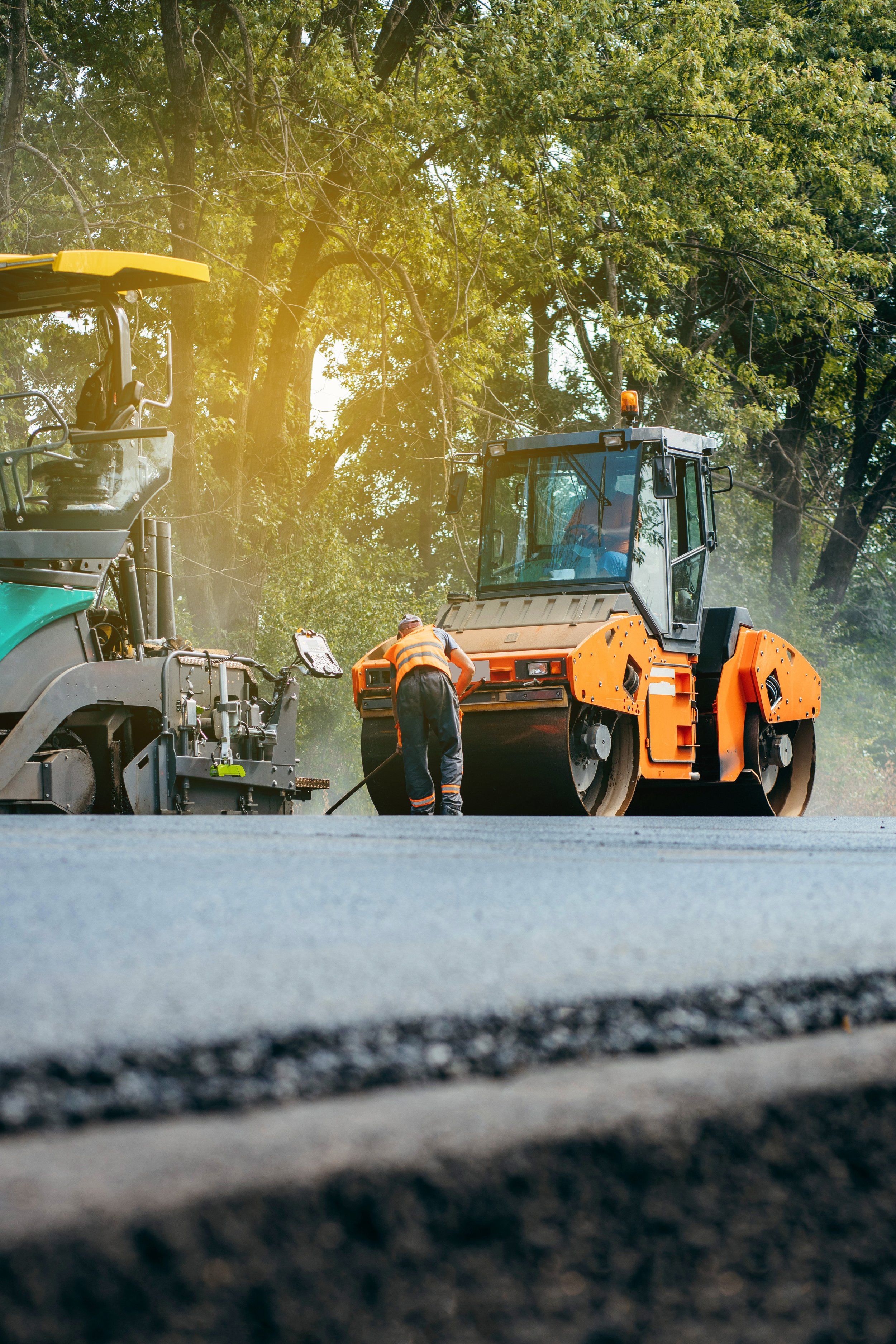 Two construction workers are working on resurfacing a road, with one smoothing freshly laid asphalt and the other operating construction equipment, surrounded by trees.