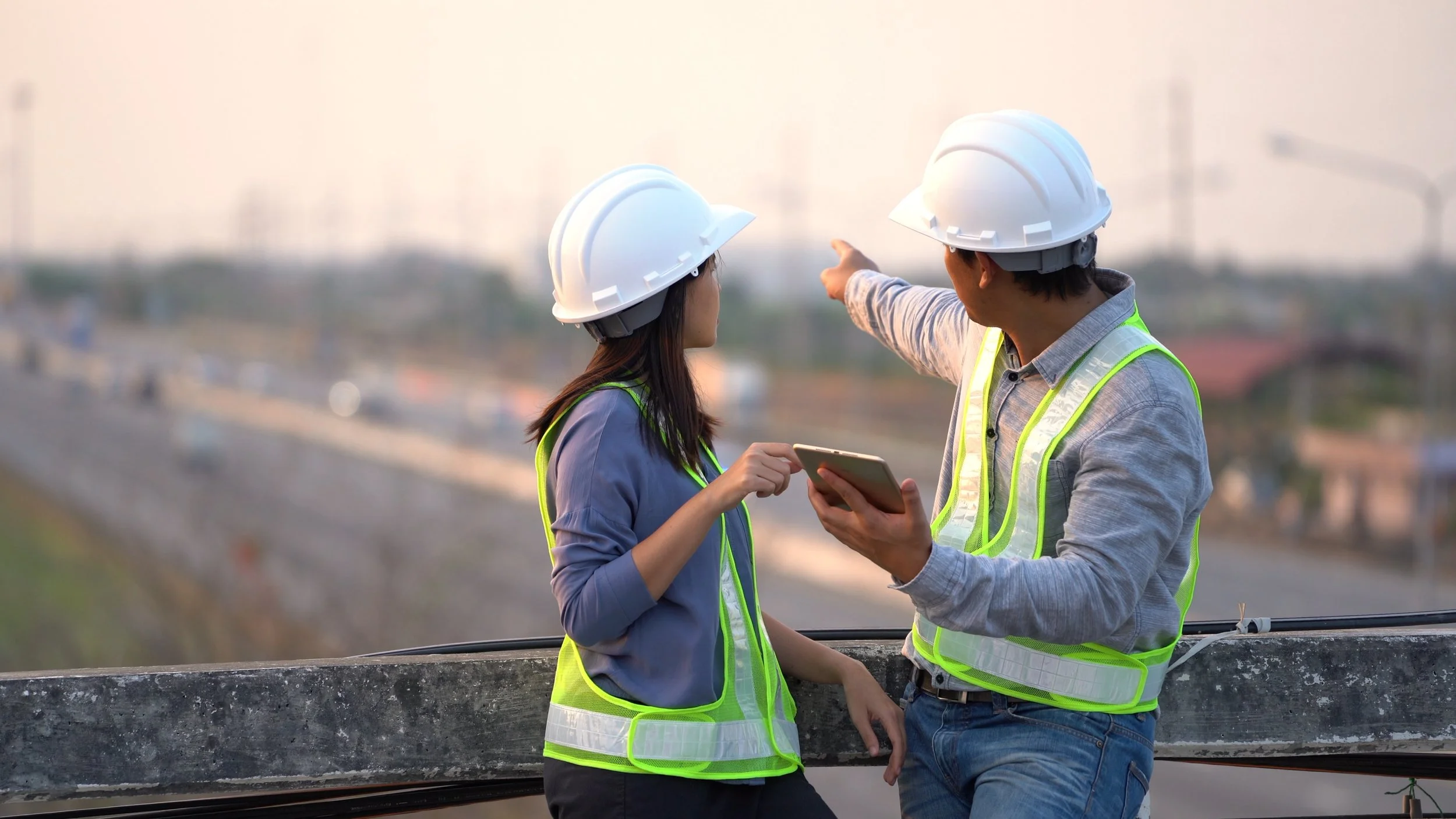 Two construction workers wearing safety helmets and reflective vests observing and discussing a construction site from an elevated platform.
