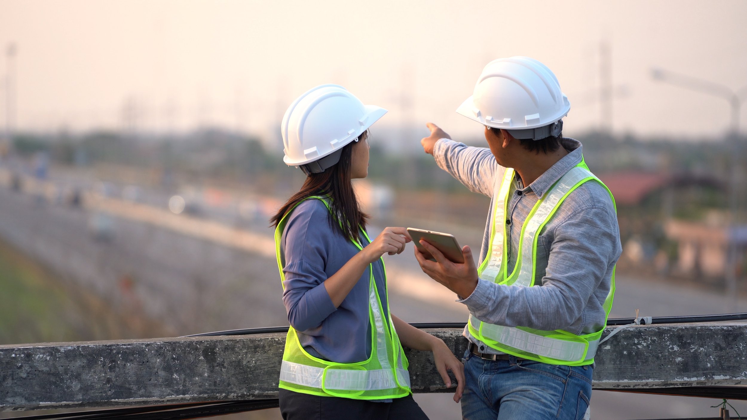 Two engineers with white hard hats and yellow reflective vests standing on a bridge overlooking a highway