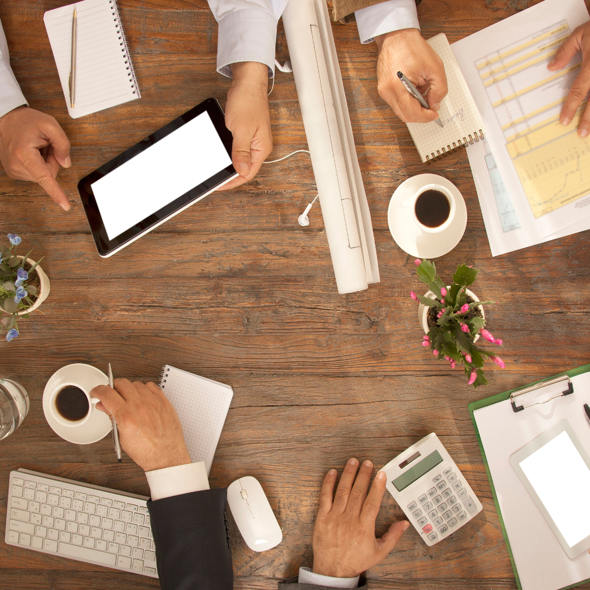 Overhead view of a cluttered wooden desk with multiple hands using a smartphone, writing notes, and working with documents, cups of coffee, a calculator, a keyboard, and potted plants.