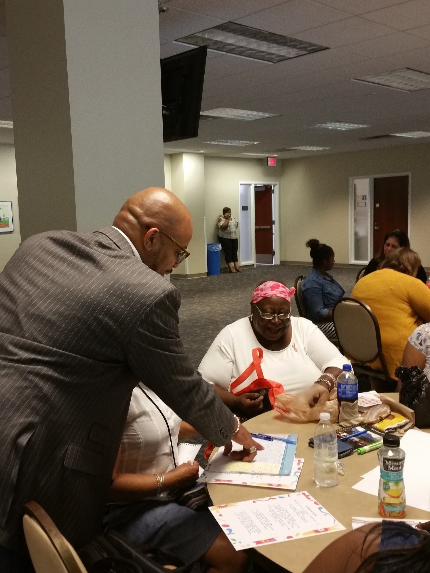 A group of people seated around a table in a conference room, with some looking at papers and a man in a suit discussing with women. There are bottles of water on the table.