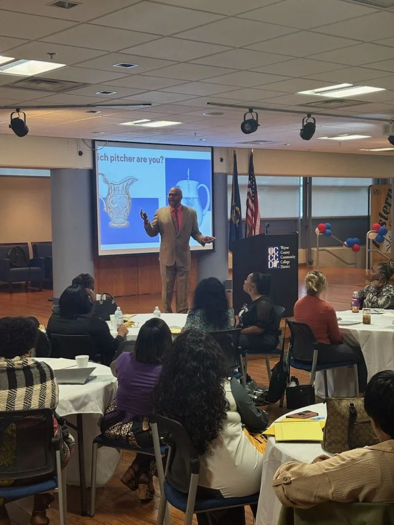 A man in a beige suit giving a presentation to an audience in a conference room. There are round tables with seated attendees, a screen behind the speaker showing two images of pitchers, and flags near the stage.