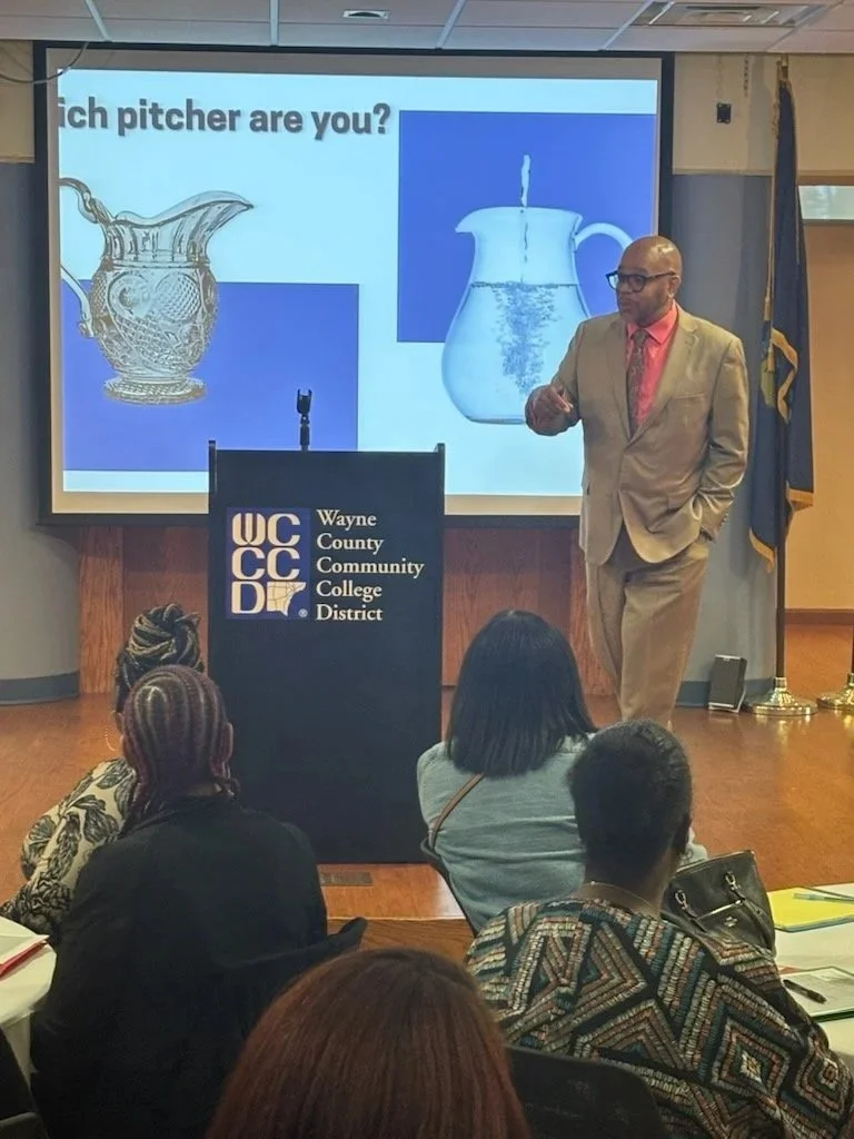 A man in a tan suit and pink shirt giving a presentation at Wayne County Community College District. He is standing next to a podium with the college's logo, facing an audience. The presentation slide behind him asks, "Which pitcher are you?" and shows images of a silver pitcher and a glass pitcher with water. Several people are sitting in the audience, watching him.