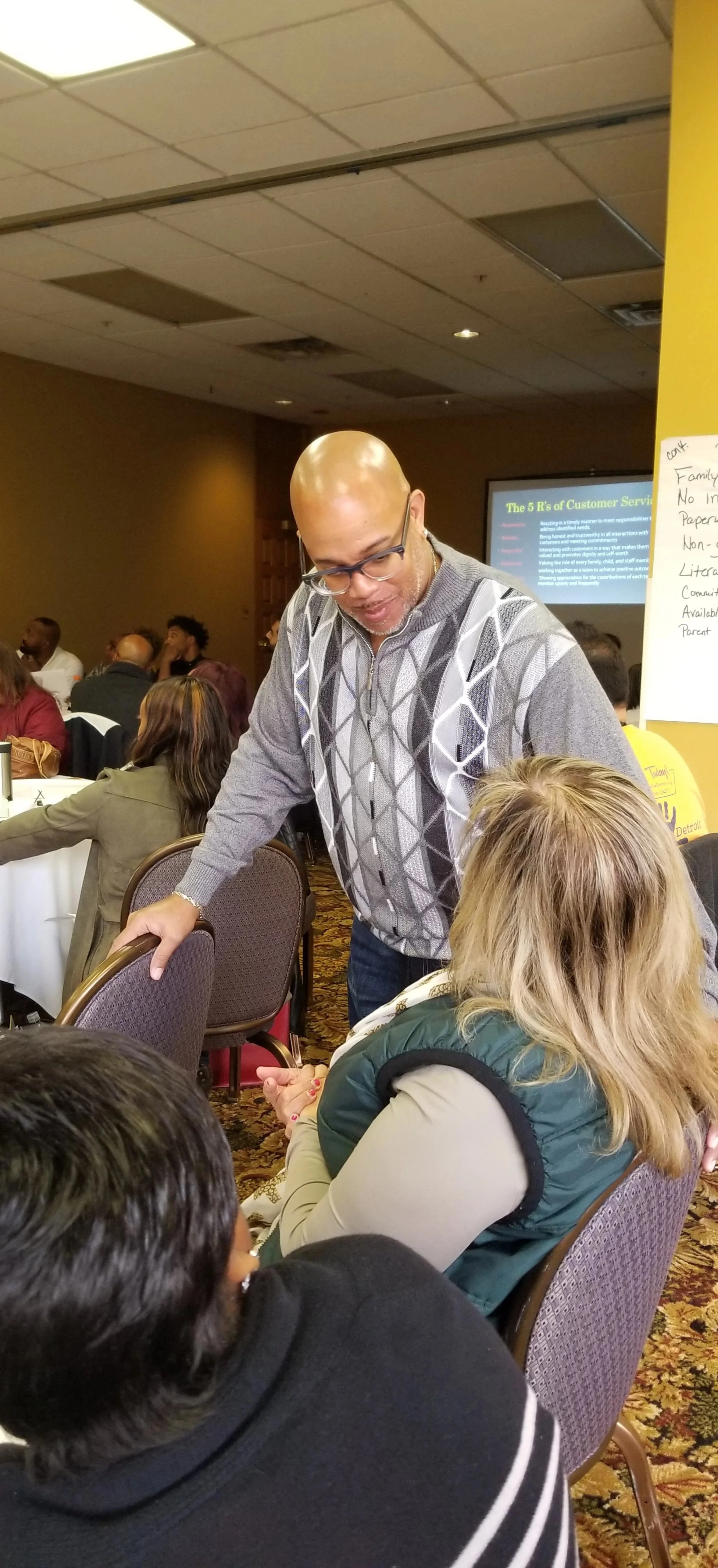A man leaning over a seated woman engaging in conversation at a conference or seminar, with other attendees visible in the background.