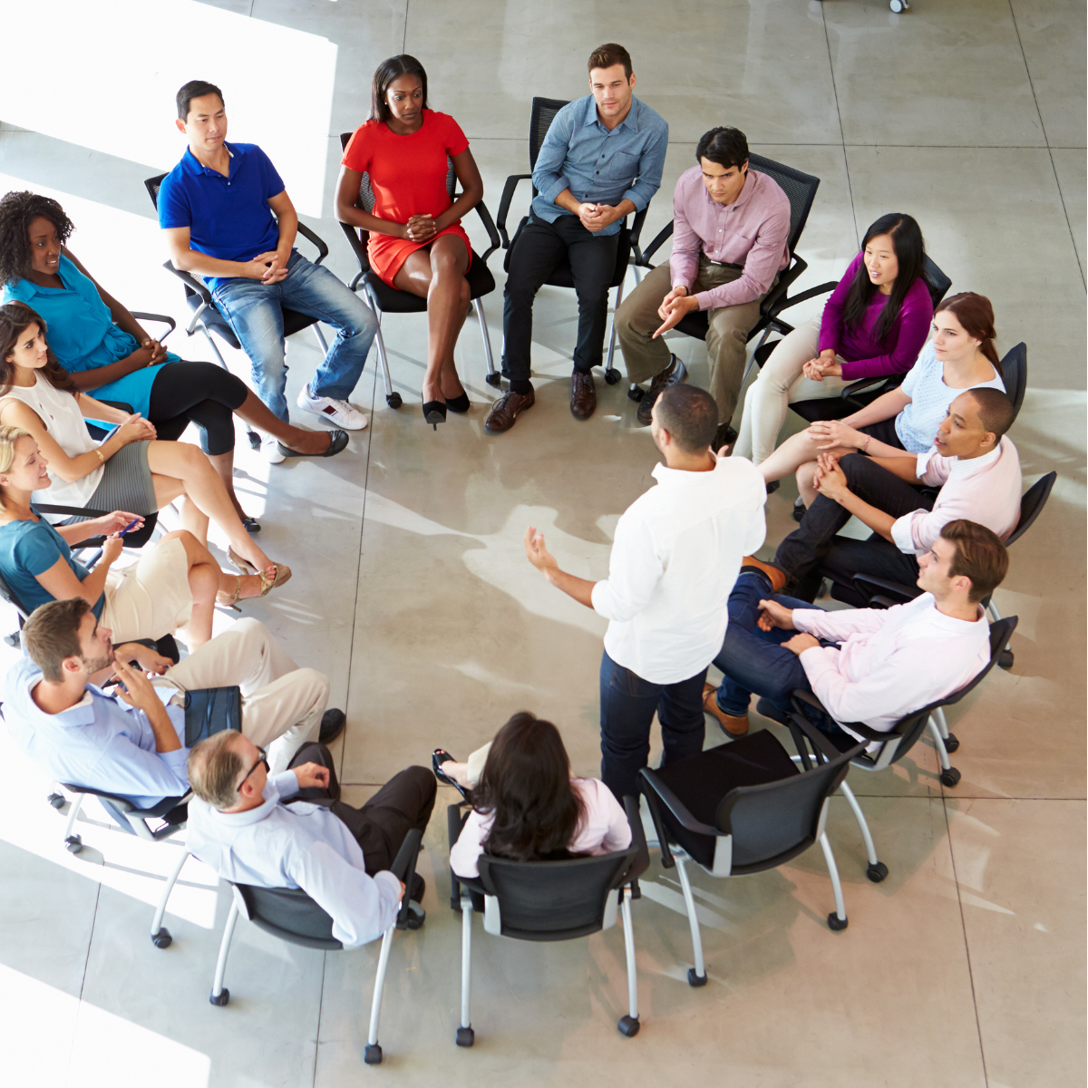 A group of people seated in a circle, listening to a person standing and speaking during a meeting or discussion in a modern space.