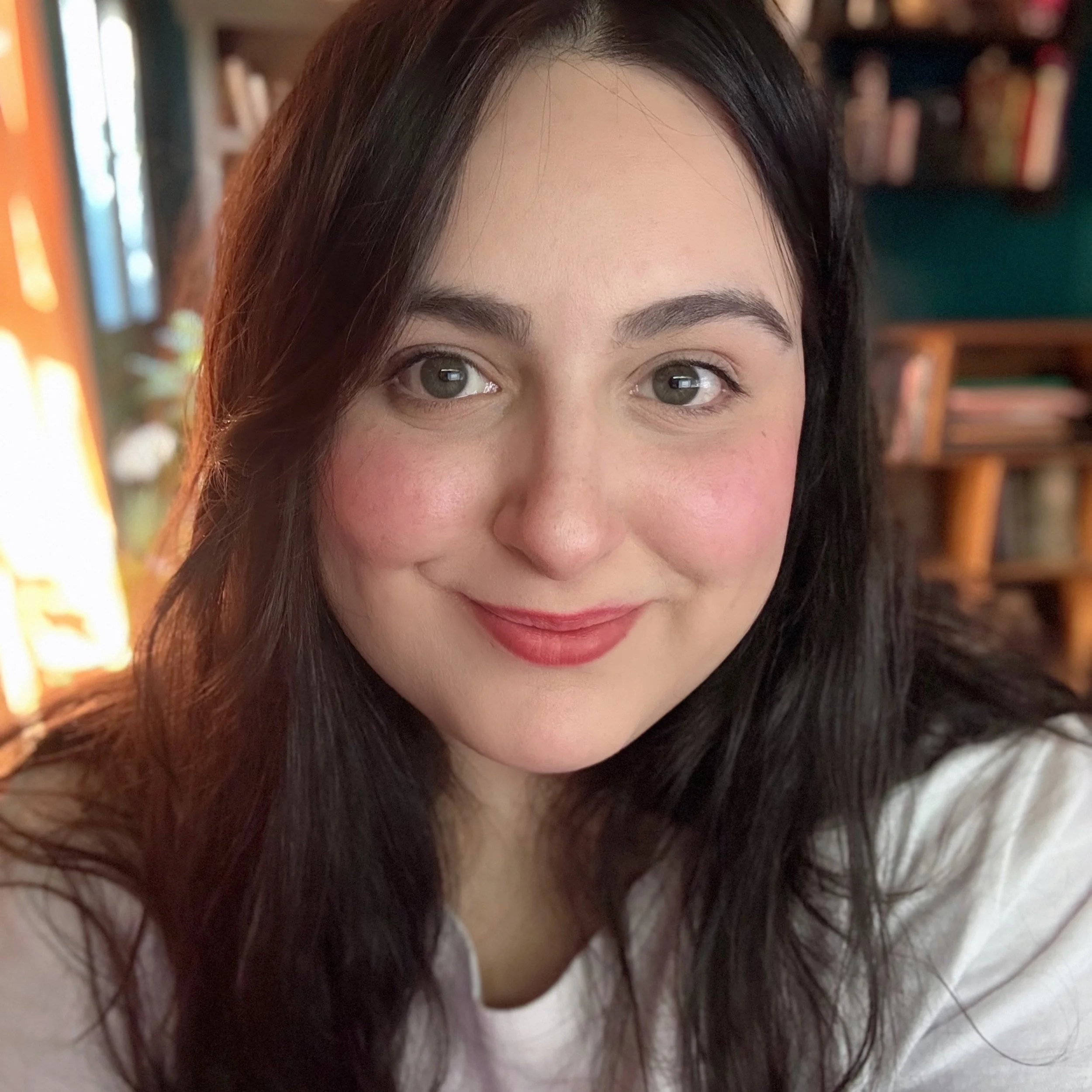 Una mujer joven con cabello oscuro y rizado, sonriendo frente a la cámara en un ambiente interior con libros en los estantes.