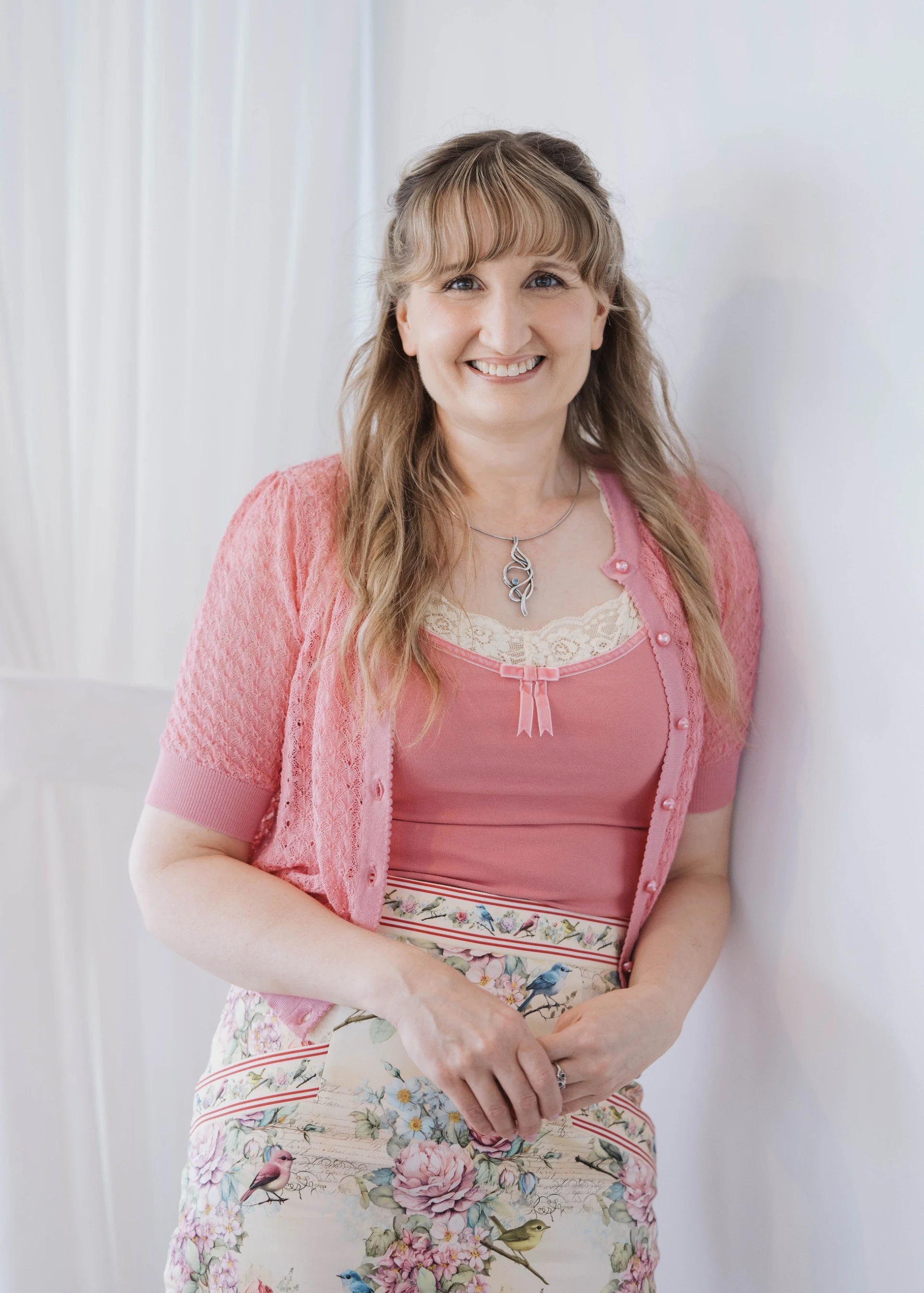 A woman with long light brown hair in loose waves, wearing a pink cardigan, a pink top with lace and a small bow, and a floral skirt with birds, standing against a white wall and smiling.