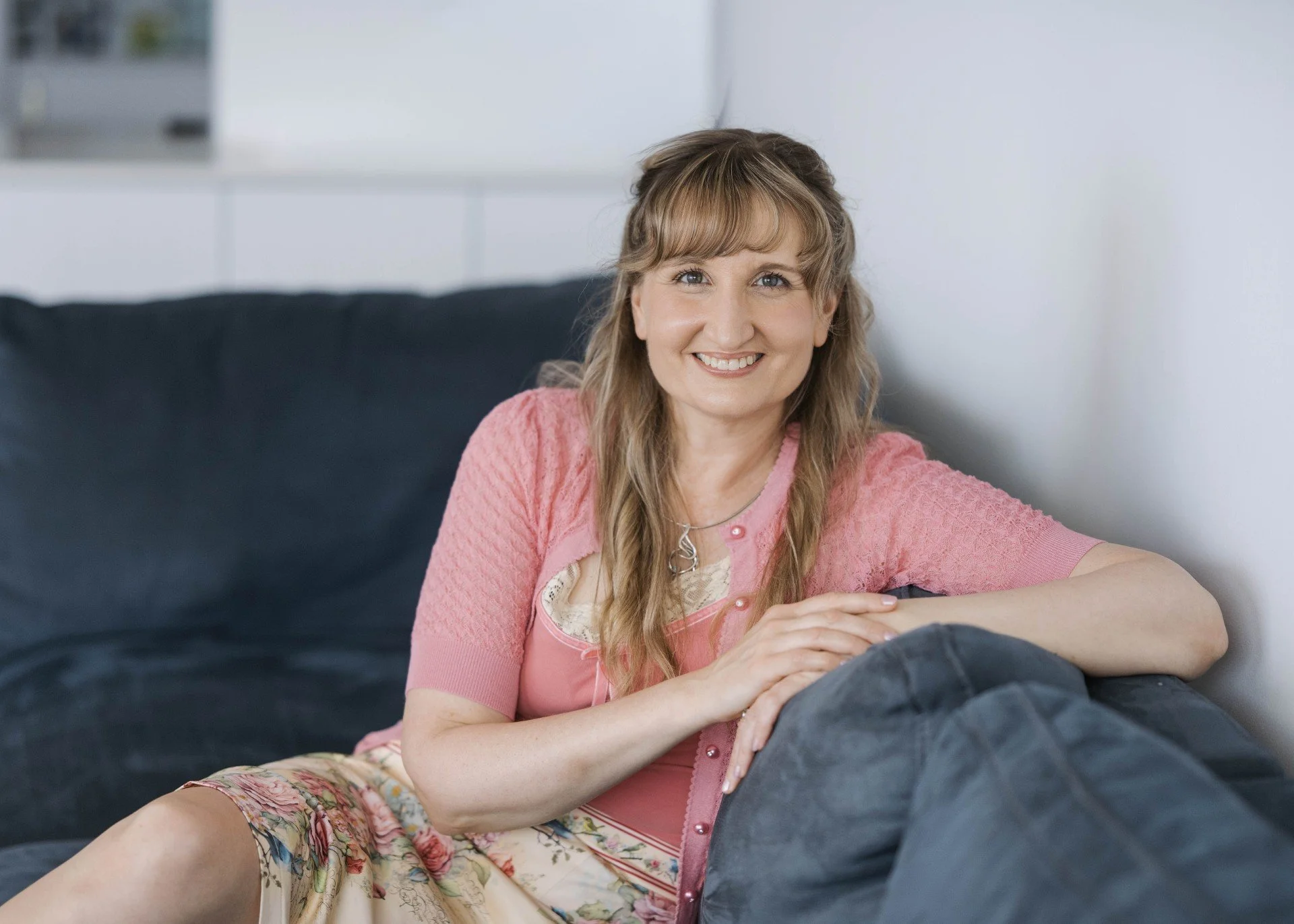 A woman with long, wavy light brown hair, wearing a pink shirt and floral skirt, smiling and sitting on a black couch in a bright room.