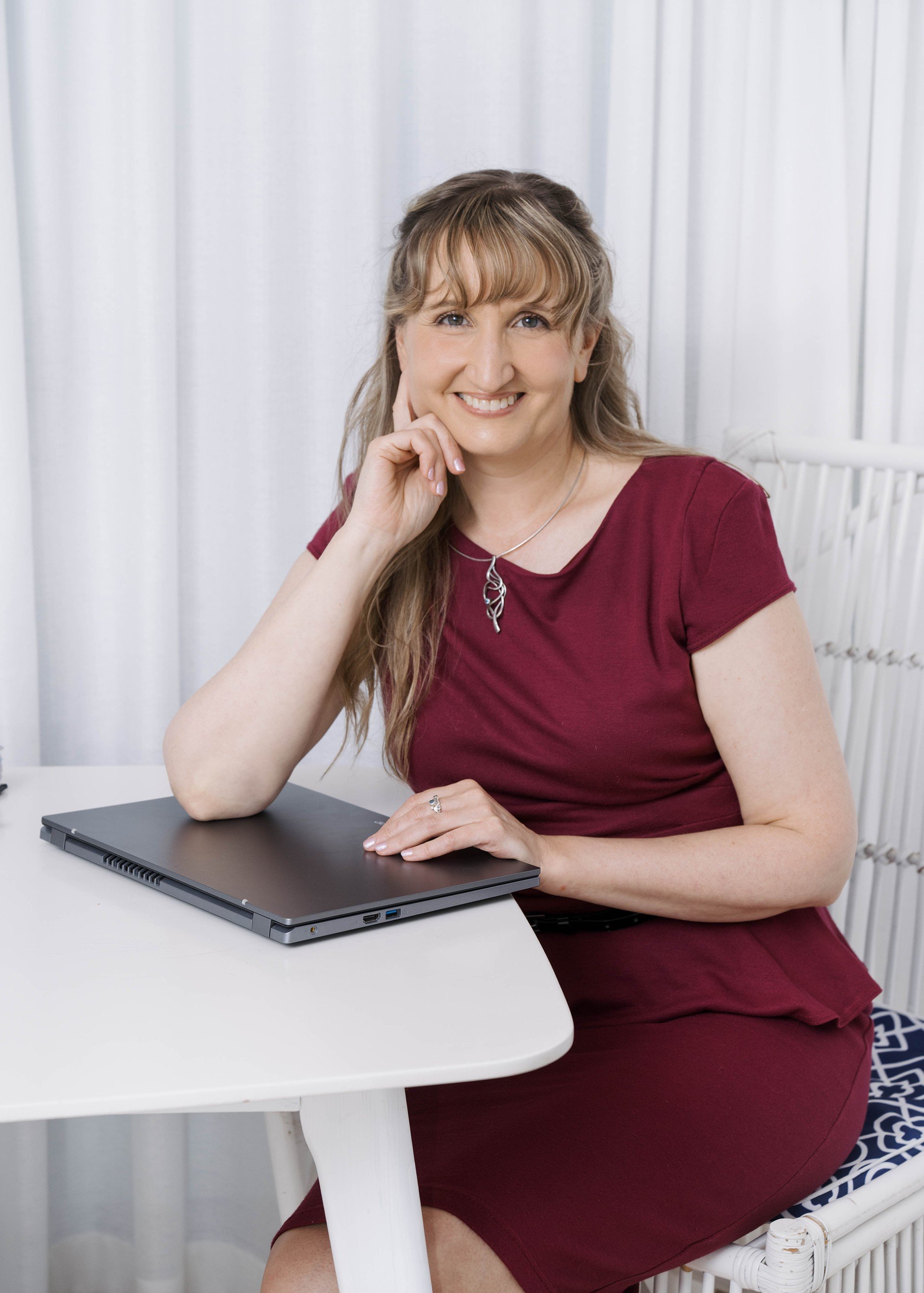 A woman with long, light brown hair sitting at a white table with a closed black laptop, smiling, wearing a burgundy dress and a silver necklace, in front of white curtains.