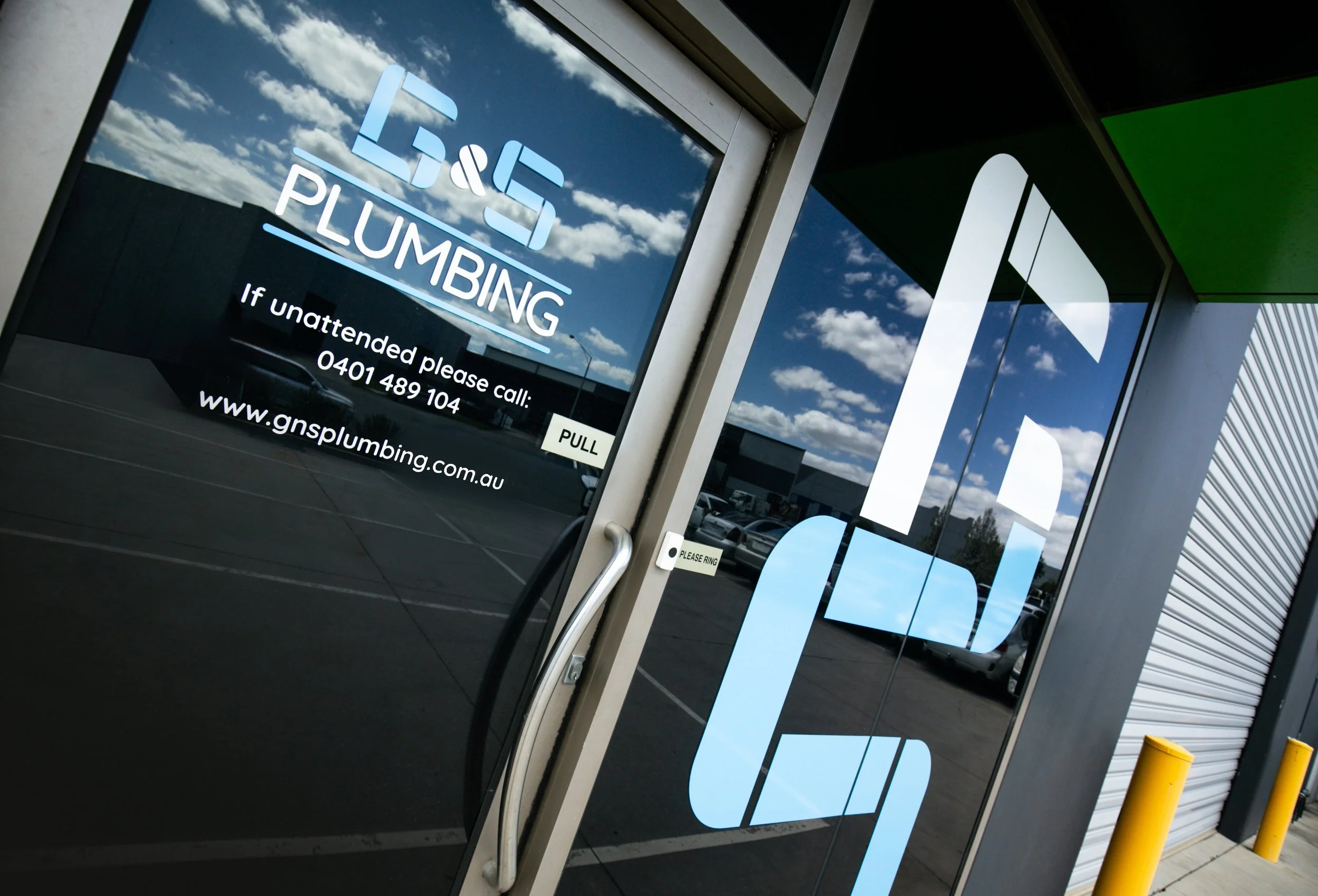 Glass door with signage for G&S Plumbing. The sign includes a phone number, website, and a note to call if unattended. The reflection shows a parking lot and a partly cloudy sky.