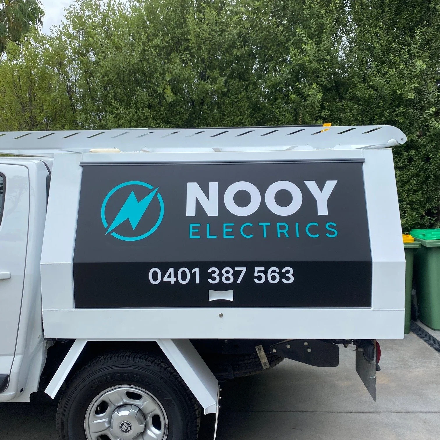 A white utility truck with a large black sign on the side that reads 'NOOY ELECTRICS,' includes a blue lightning bolt logo, and a phone number '0401 387 563.' The truck is parked on a driveway with trees in the background and green garbage bins nearb