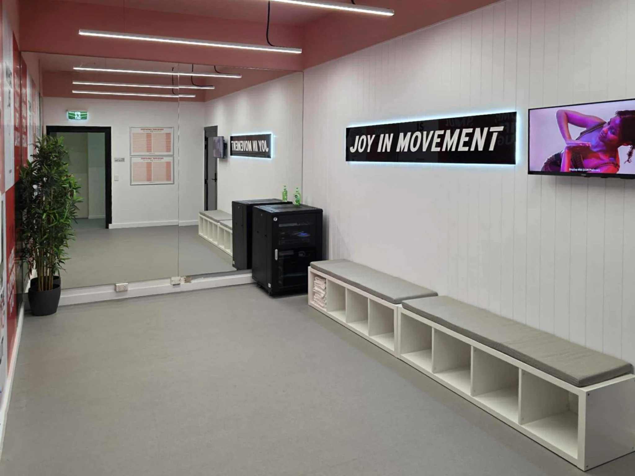 An interior gym lobby with a white wall, benches, potted plant, and digital screens. One screen displays a woman exercising, another shows the text 'Joy in Movement'.