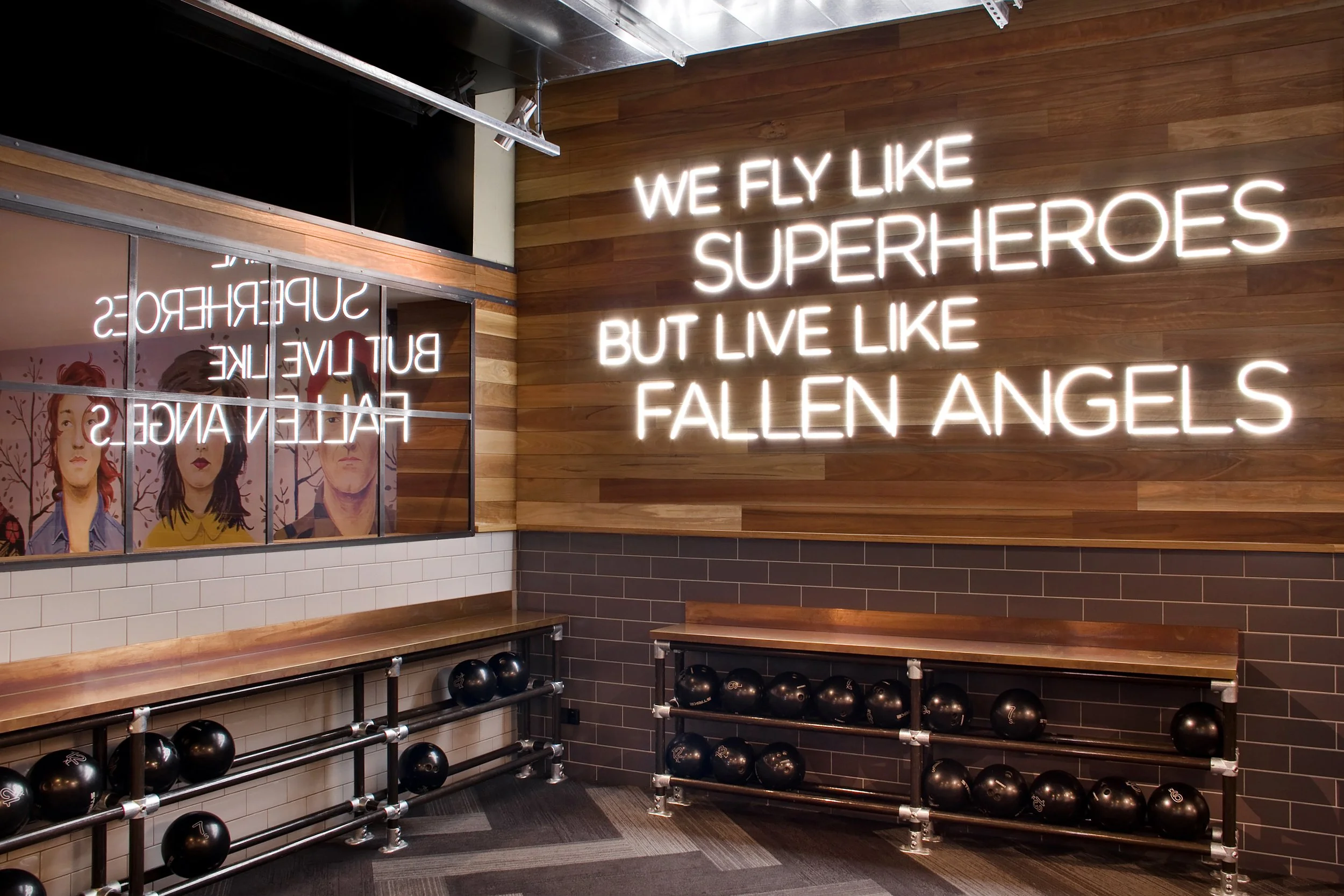 Neon sign with the quote 'We fly like superheroes but live like fallen angels' on a wooden paneled wall in a fitness or gym studio. Below, there are black kettlebells on metal racks and a mirror reflecting illustrated portraits of women.