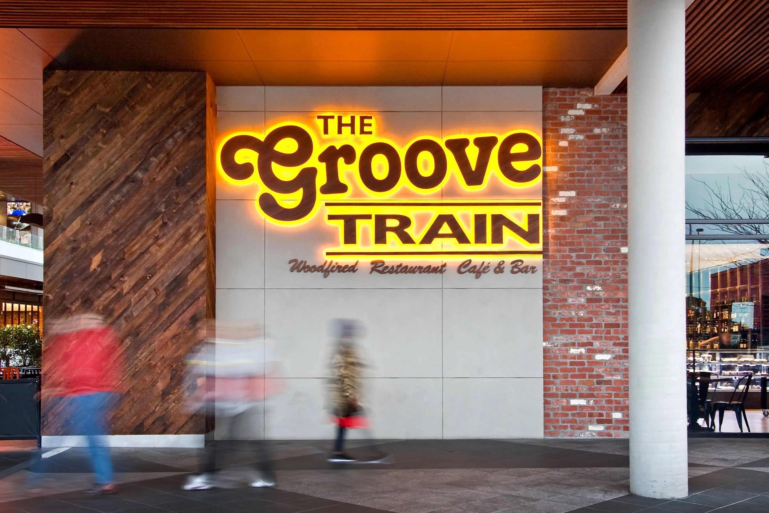 Neon sign reading 'The Groove Train' with subtext 'Woodfired Restaurant Café & Bar' on a wall outside a restaurant, with blurred people walking past.