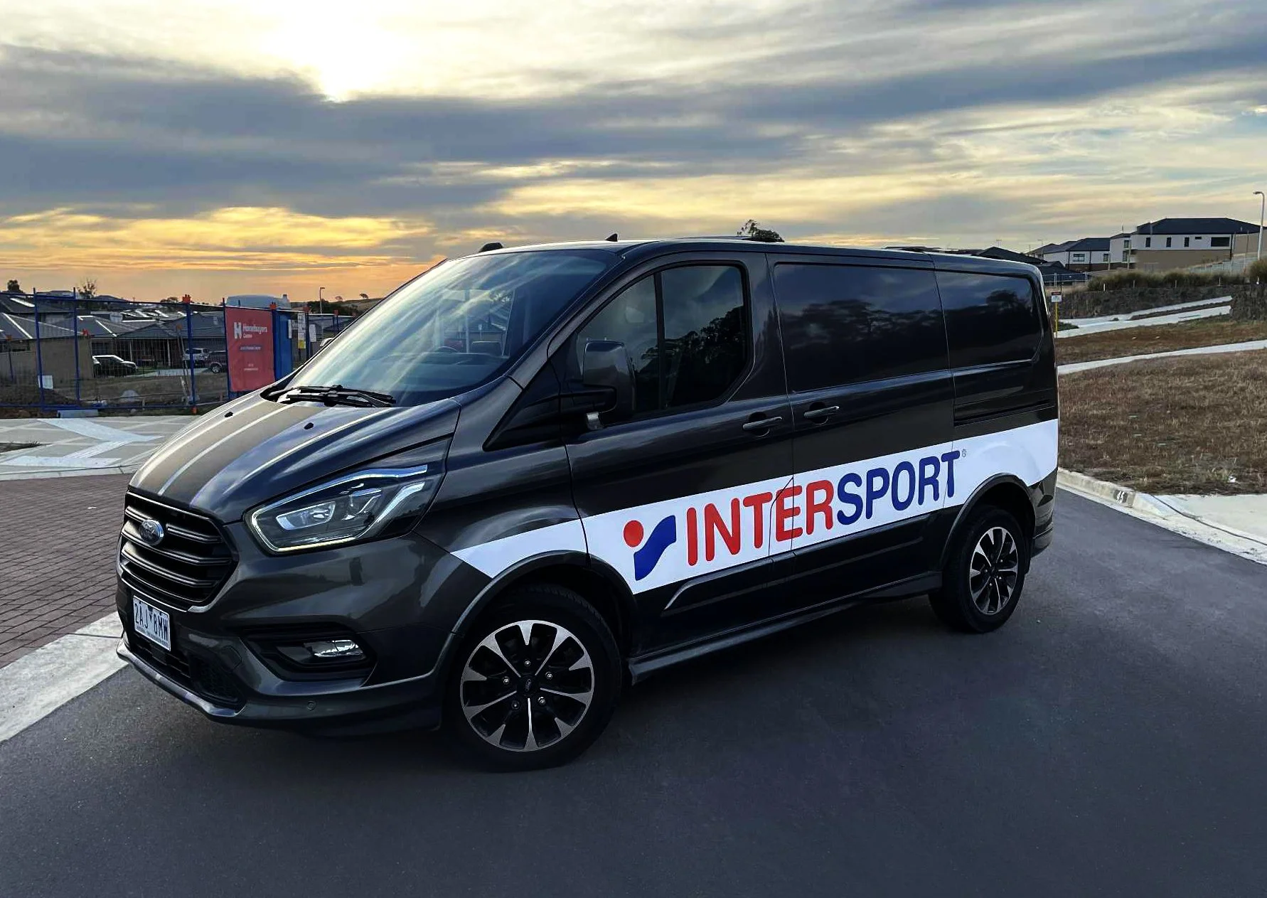 Black van with 'INTERSPORT' logo parked on the side of the road during sunset, with residential and commercial buildings in the background.