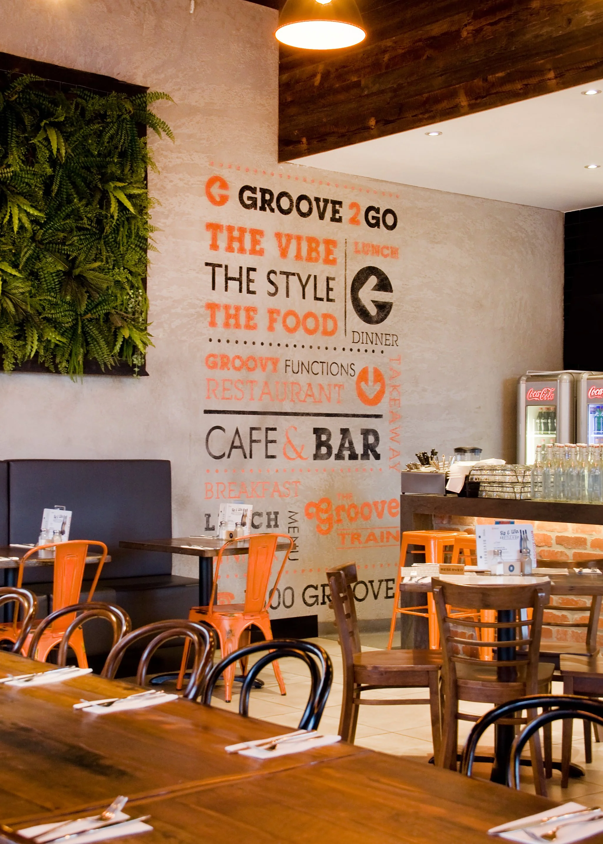 Interior of a restaurant with a wall decorated with a large design featuring words like 'Groove 2 Go,' 'The Vibe,' 'The Style,' 'The Food,' and 'Cafe & Bar.' There are tables and chairs, including orange metal chairs, wooden chairs, a large wooden ta