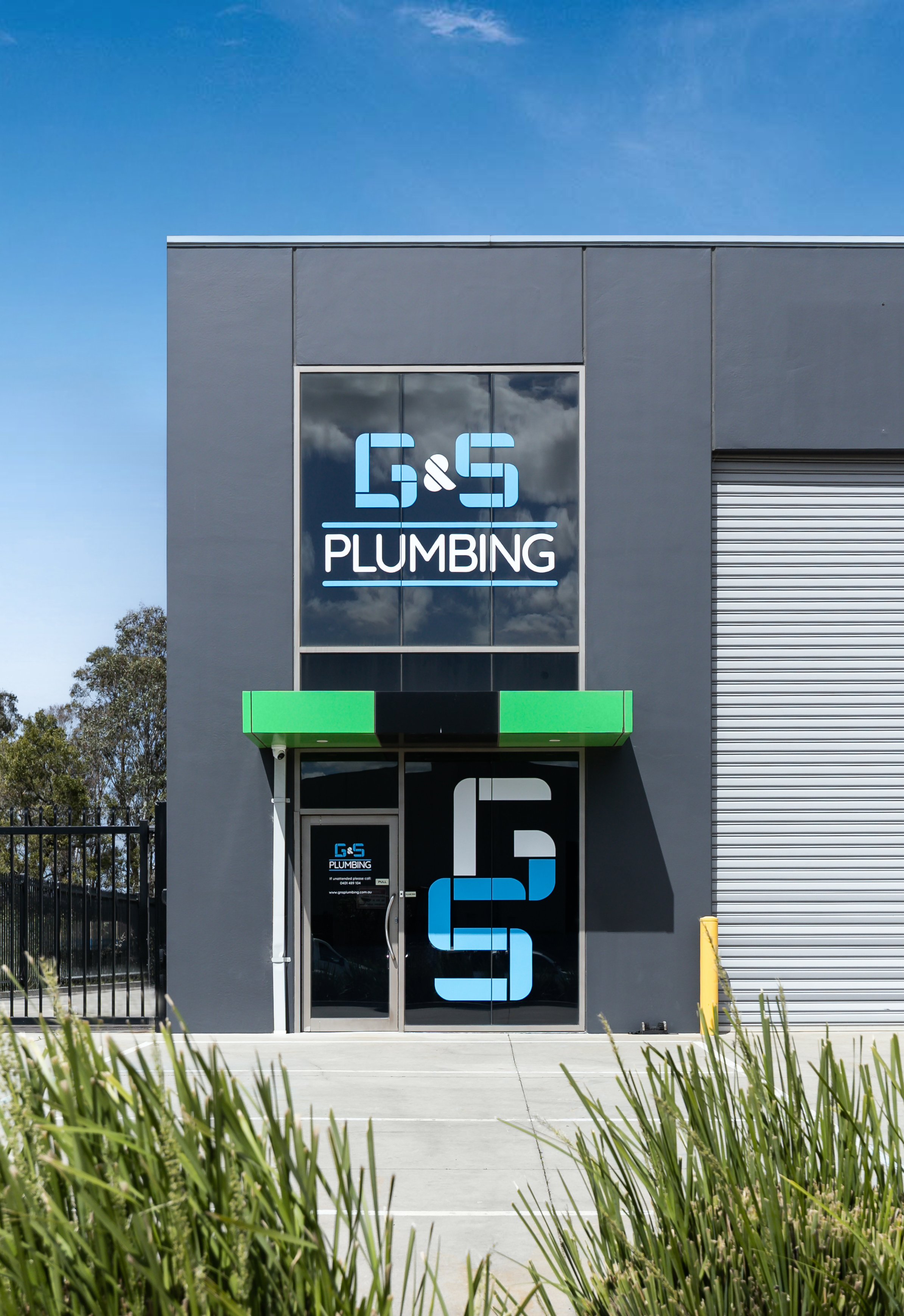 Exterior view of G & S Plumbing business with gray building, glass door, and signage, under a blue sky with some clouds.