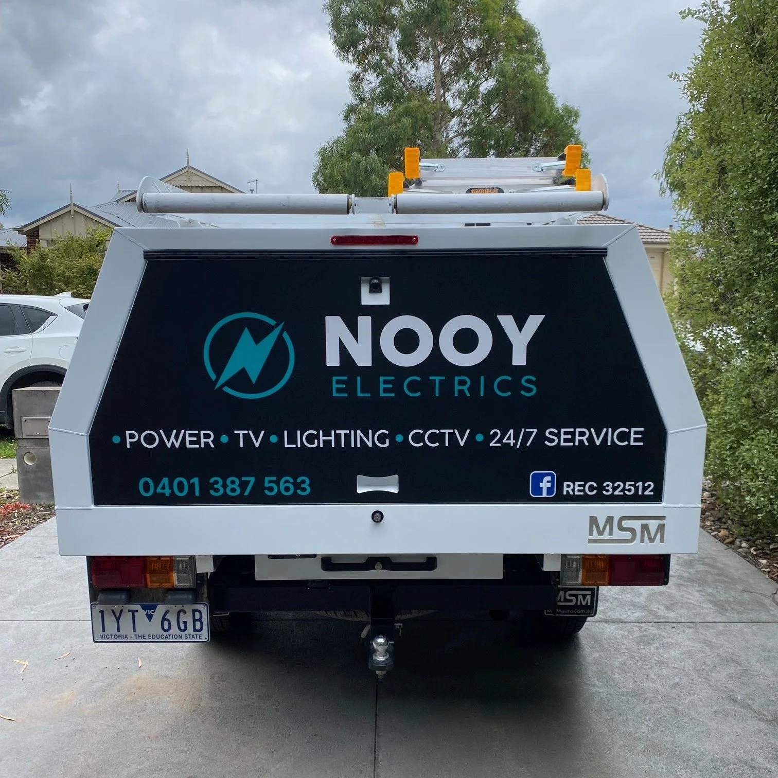 Rear view of a service vehicle with advertising for NOOY Electrics, a business offering power, TV, lighting, CCTV, and 24/7 service. The vehicle has a ladder on top and the license plate reads 1YT 6GB.