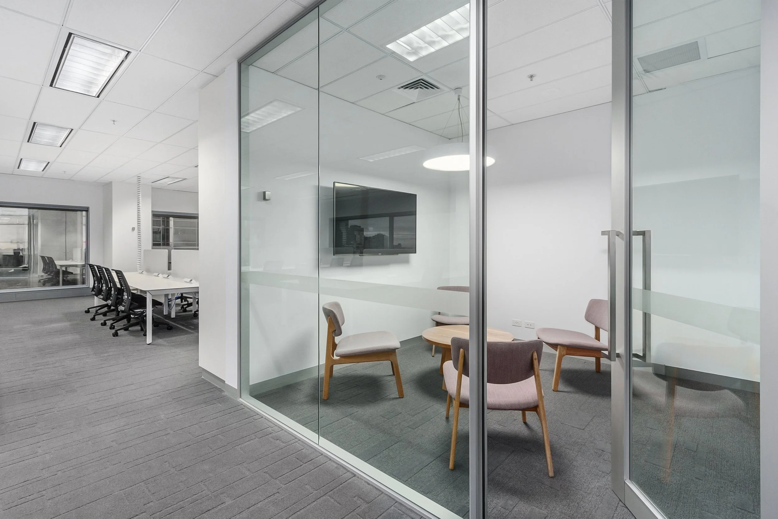 Modern office conference room with glass walls, a round table, and pastel-colored chairs, adjacent to a larger meeting space with a long table and black chairs.
