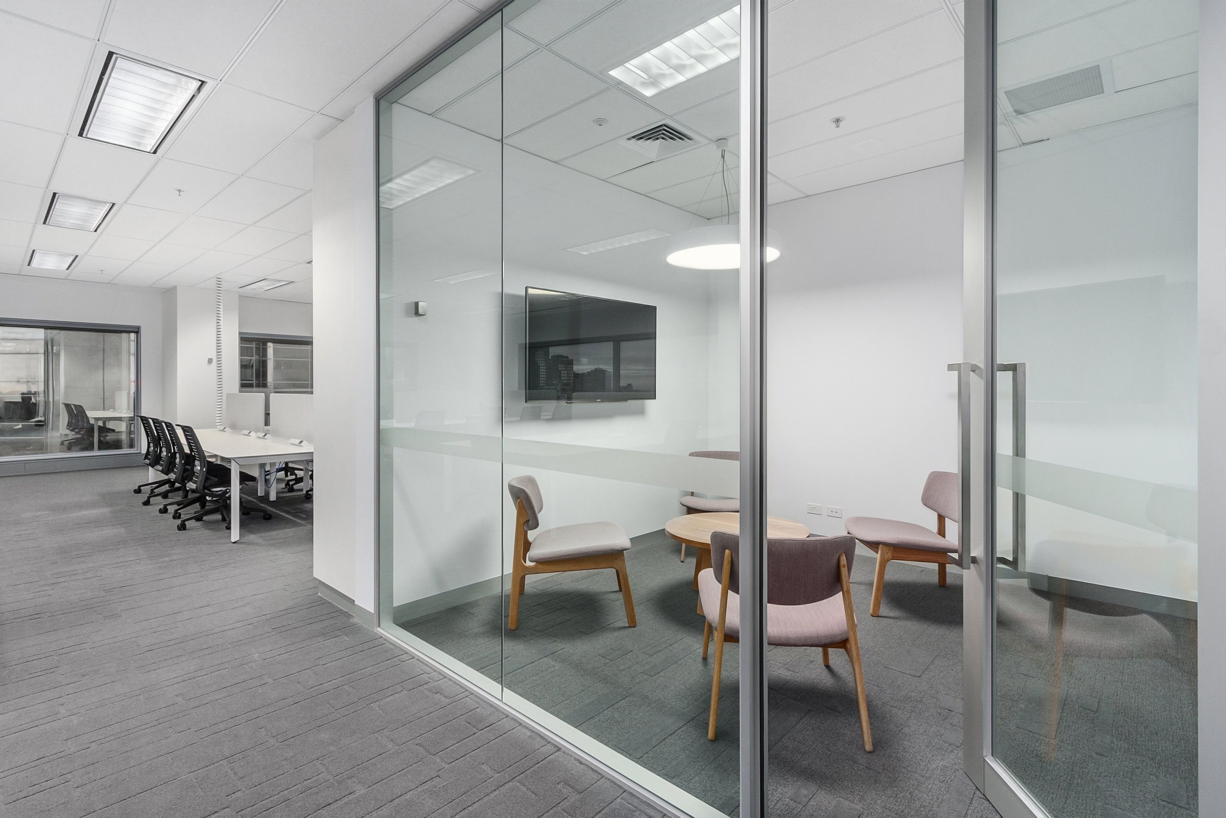 Modern office conference room with glass walls, a round table, and pastel-colored chairs, adjacent to a larger meeting space with a long table and black chairs.