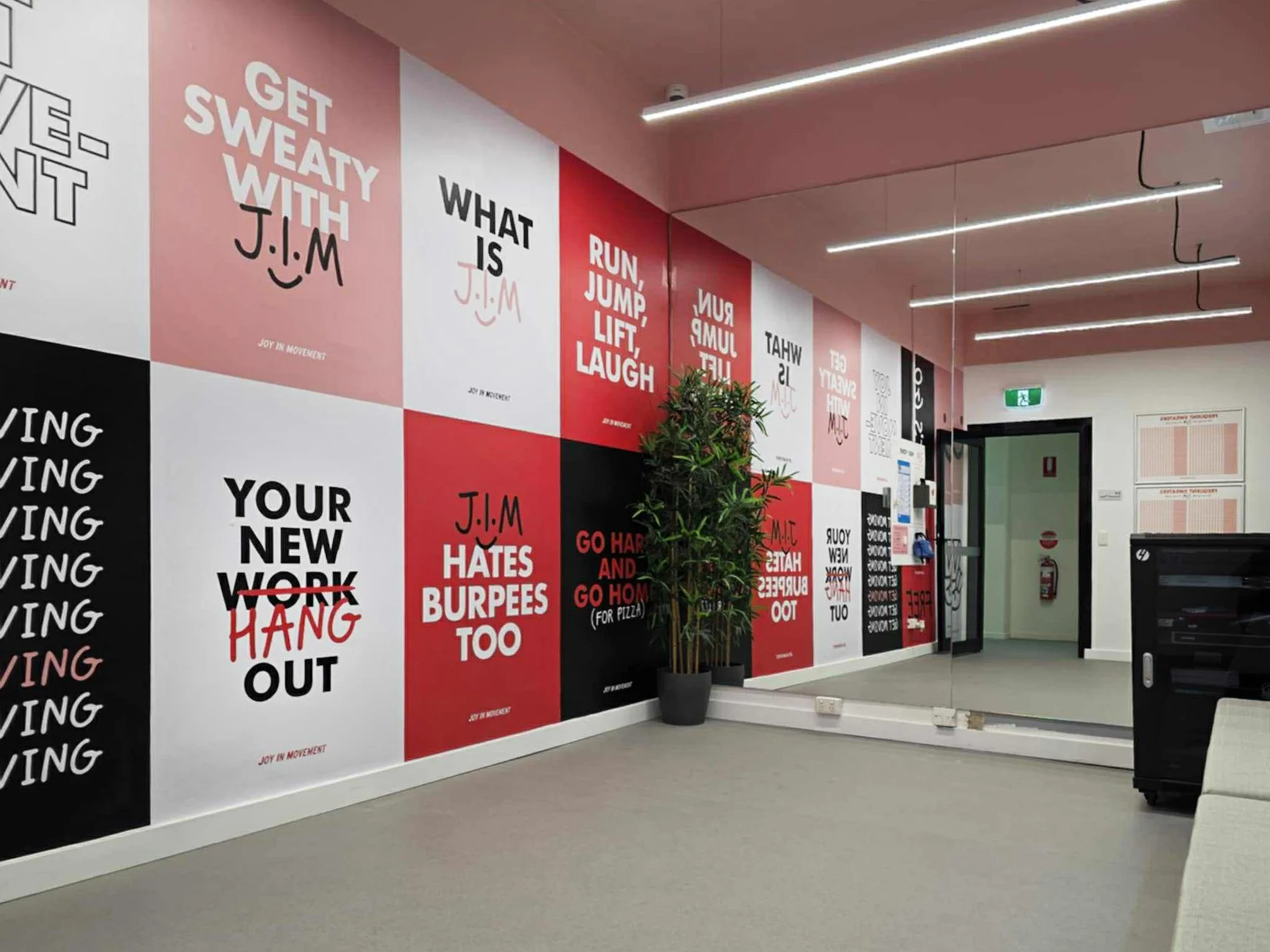 Office wall decorated with pink, red, black, and white motivational posters with quotes and phrases, a potted plant, a mirror, a fire extinguisher, and a door.