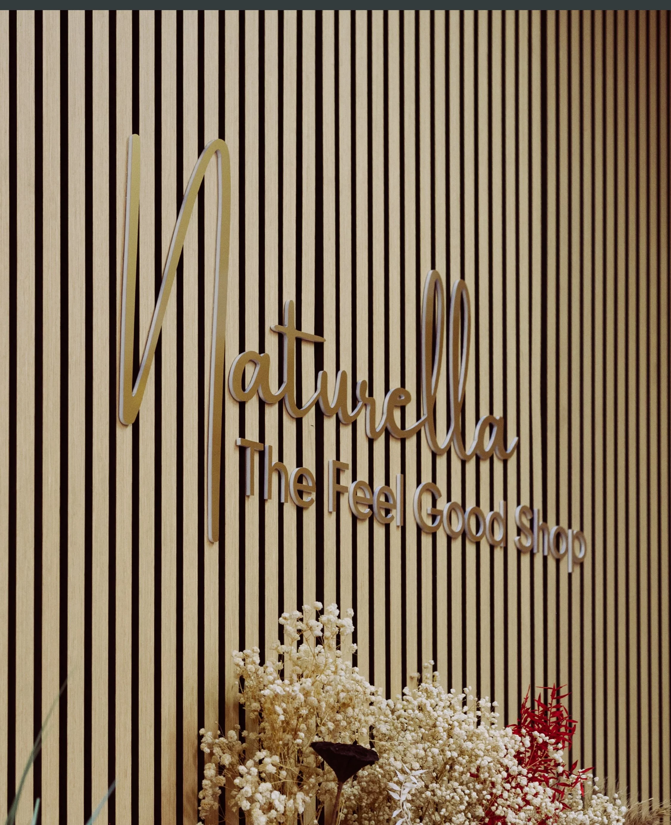 Wooden wall with a sign that says "Naturella The Feel Good Shop" in gold cursive and block letters, and a bouquet of white flowers at the bottom.
