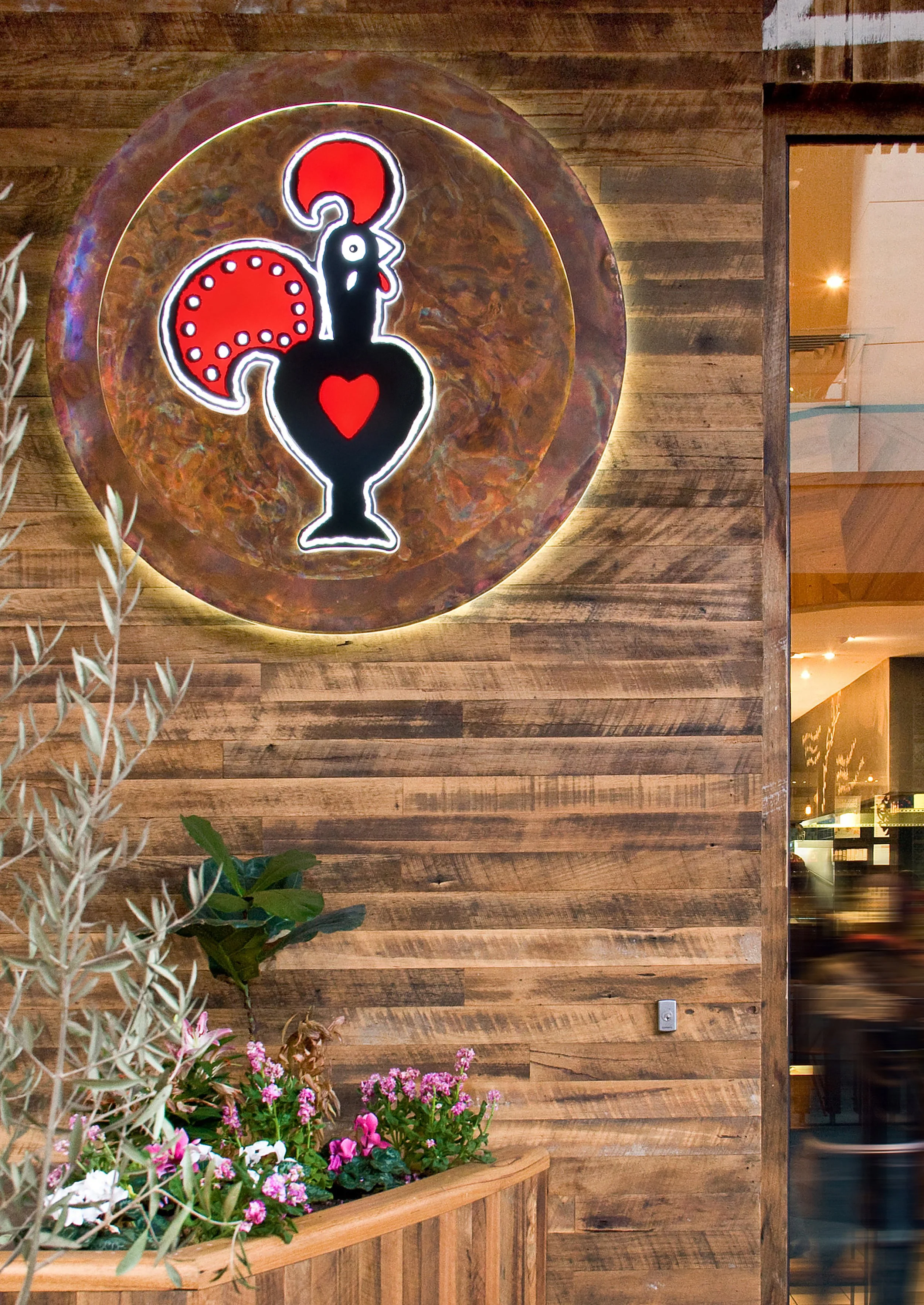 Decorative wall art with a stylized illuminated black vase with a red heart, red circle, and red dotted semicircle, mounted on a wooden wall in a restaurant or cafe, with potted pink and white flowers below.