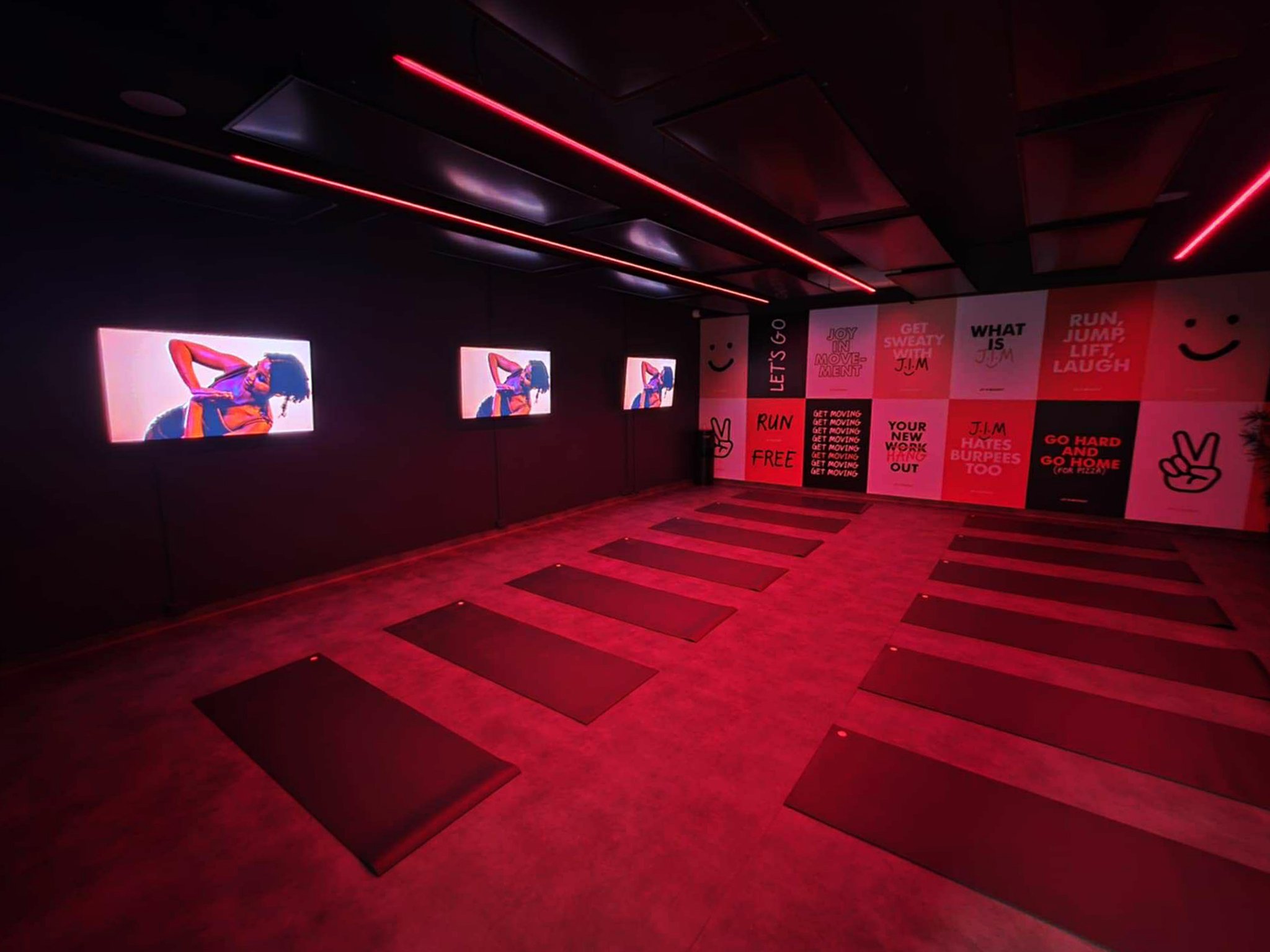 A dimly lit workout room with red neon lighting, multiple yoga mats arranged in rows, three wall-mounted screens displaying a woman stretching, and motivational posters with phrases like "Run Free" and "Get Sweaty with J.M" on the walls.