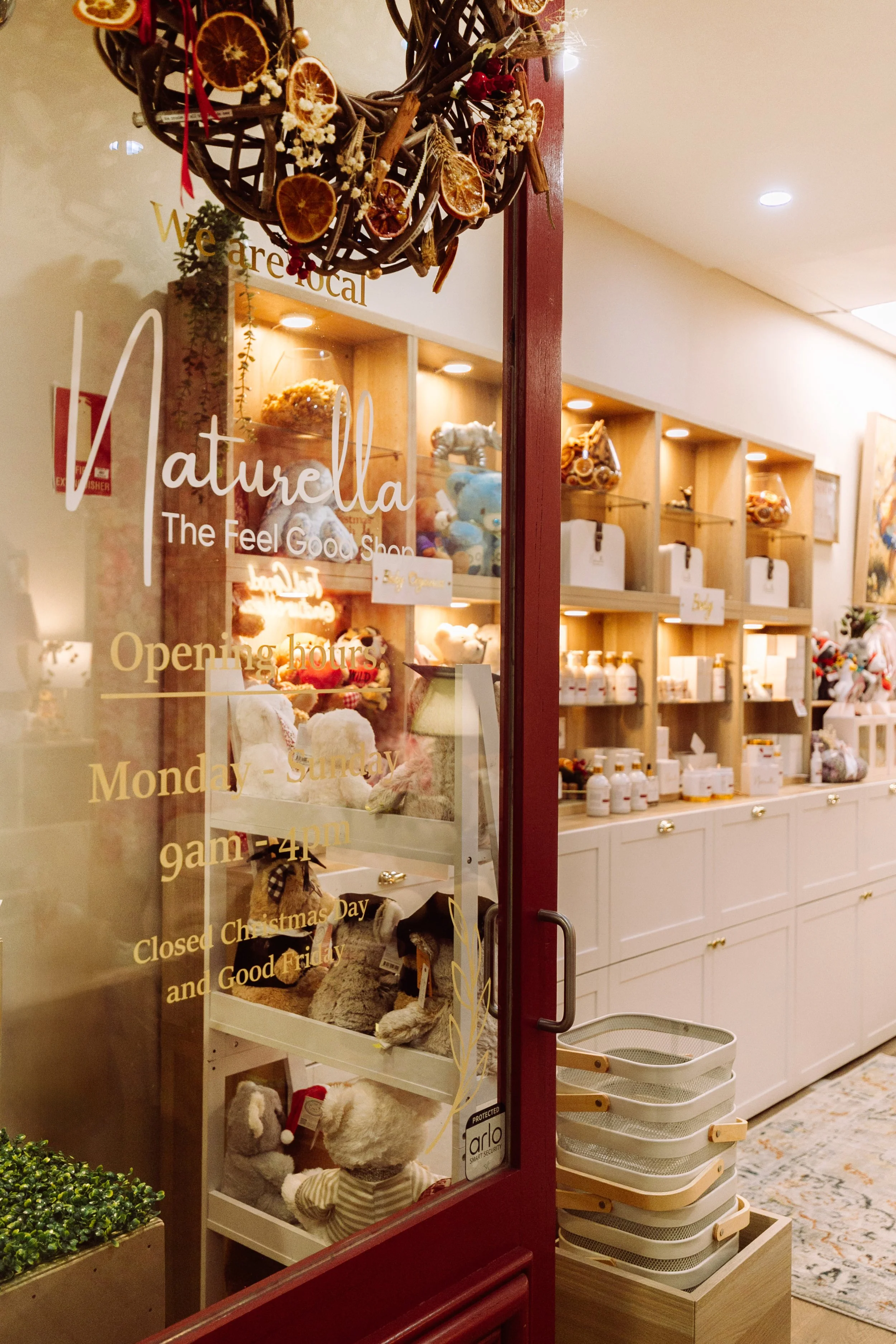 View through the glass door of Naturella, a shop that sells stuffed animals, gifts, and health products. The shop's opening hours are Monday to Sunday, 10 am to 4 pm, and it is closed on Christmas Day and Good Friday. There is a decorative wreath mad