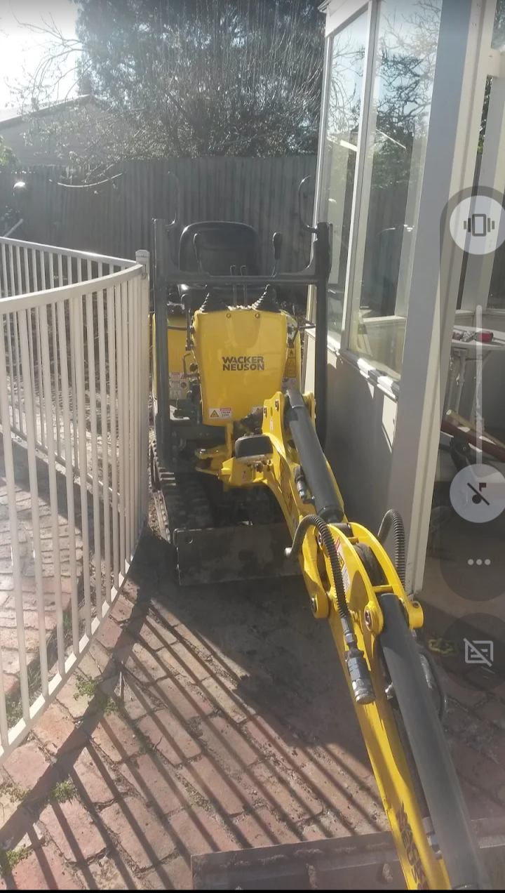 A yellow Wacker Neuson mini excavator parked outside on a brick pathway, with a glass enclosure on the right and a white metal fence on the left, casting shadows on the ground.