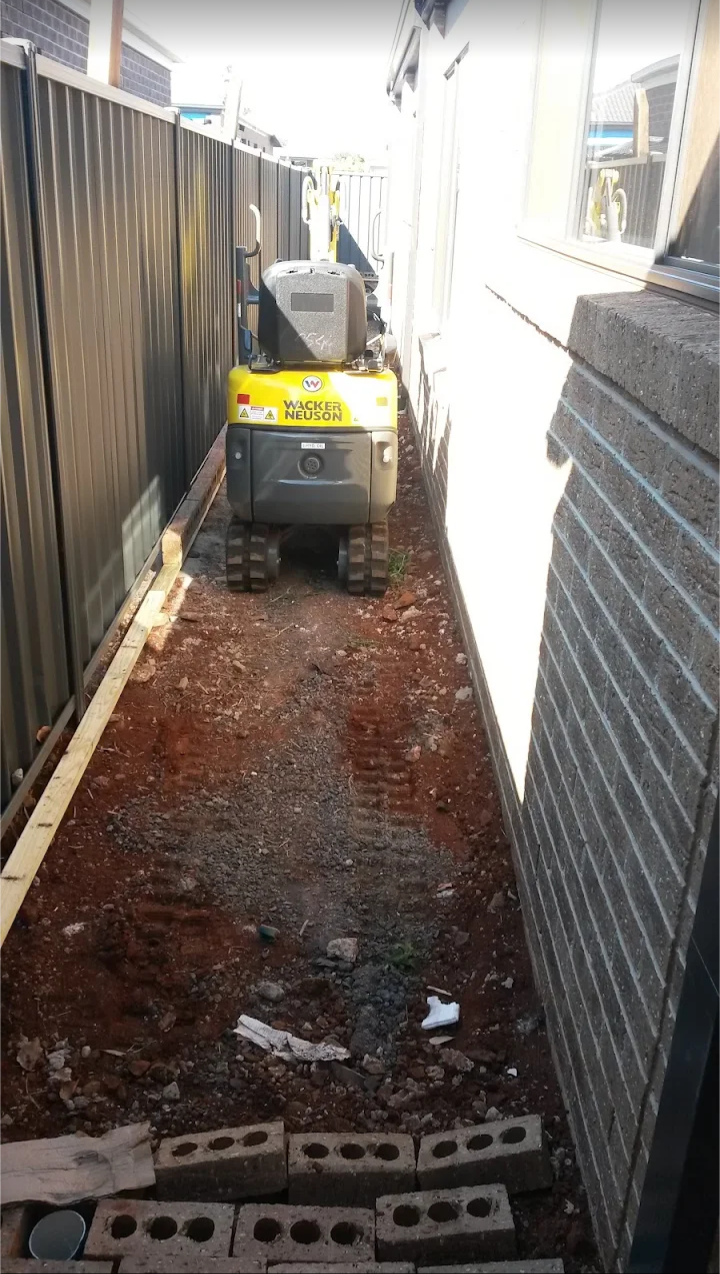 A narrow outdoor space with a yellow Wacker Neuson excavator on reddish dirt ground, bordered by a black metal fence on the left and a brick wall on the right, with construction materials and tools visible.